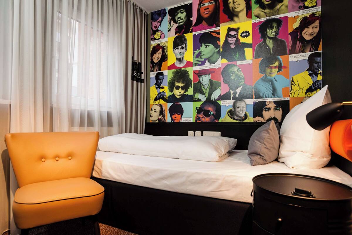 A hotel room with a bed, an orange chair, a black nightstand, and a wall with colorful posters.