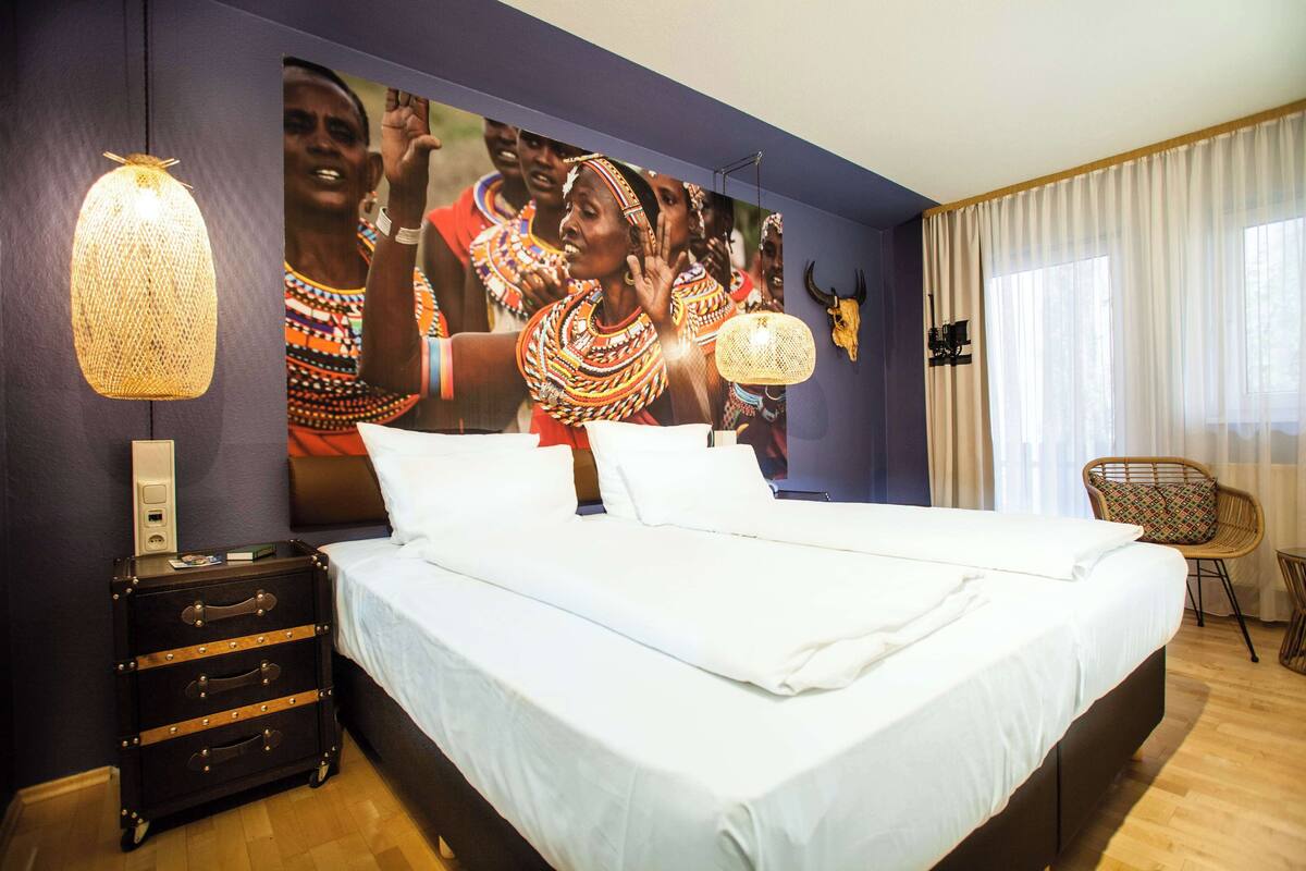 A hotel room with a large bed, a nightstand, a wall-mounted lamp, a decorative cow skull, and a window with curtains.