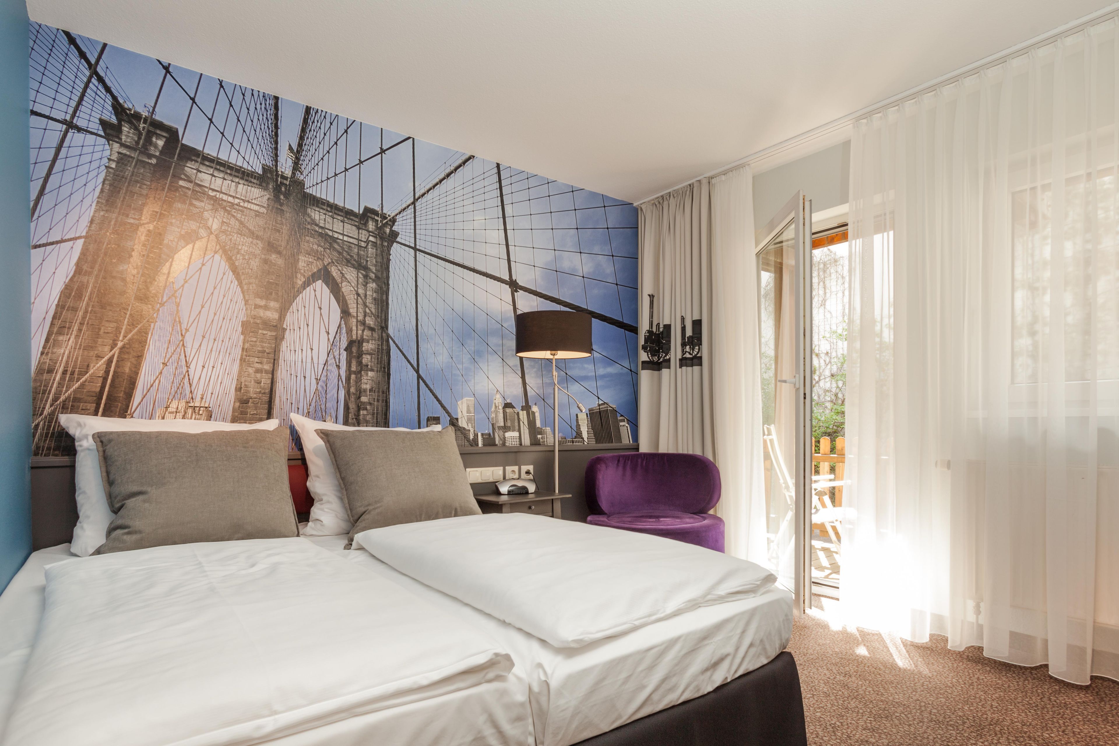 Ein Hotelzimmer mit einem Bett, einem violetten Sessel, einer Lampe und einem Wandbild einer Brücke.