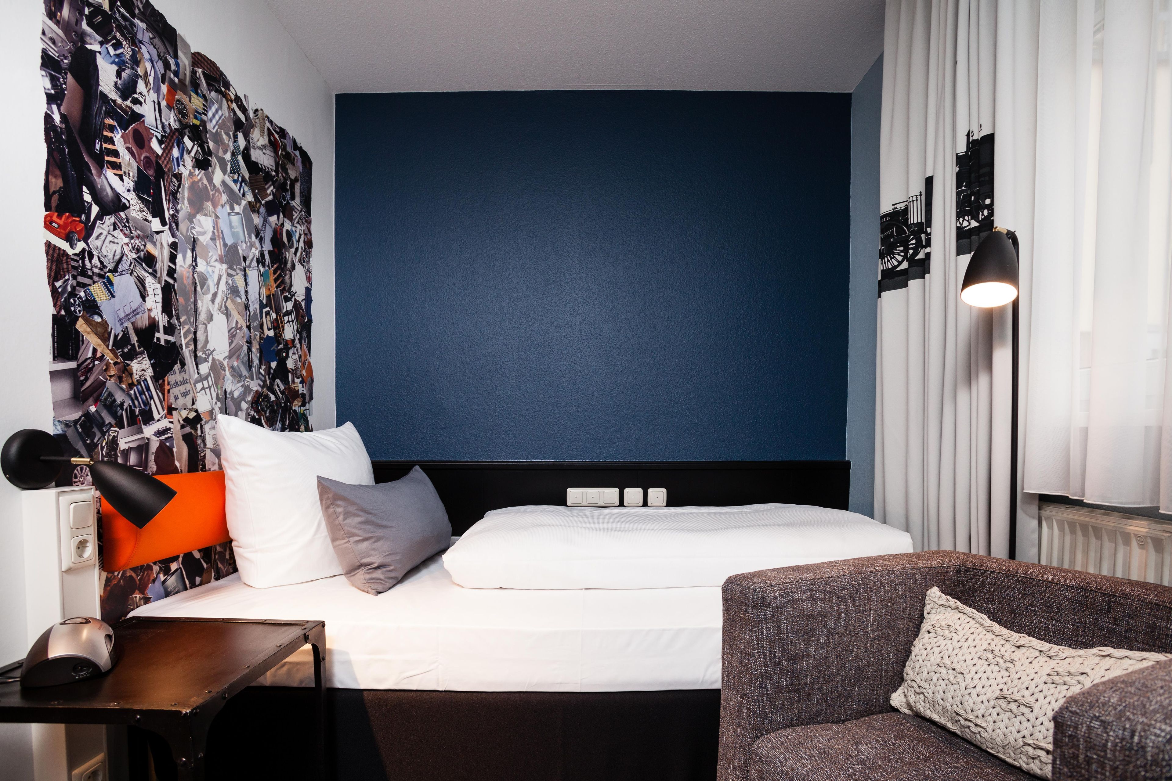 A hotel room with a large bed, a desk, a chair, a wall with a blue accent, and a curtain with a black and white pattern.