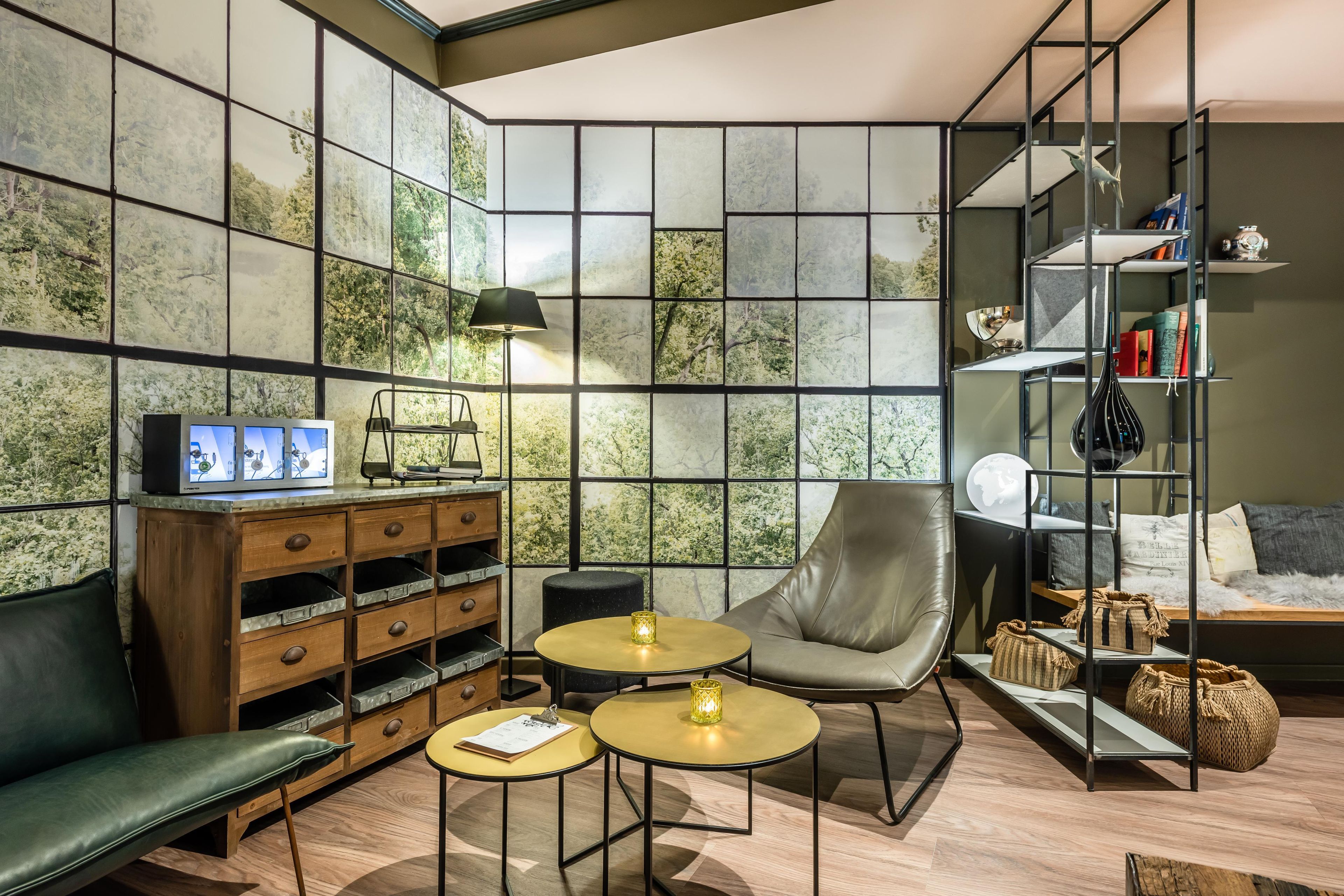 Ein modernes Wohnzimmer mit einem Ecksofa, einem Ledersessel, zwei kleinen runden Tischen, einem Bücherregal und einer Wand mit einem Wandbild im Naturdesign.