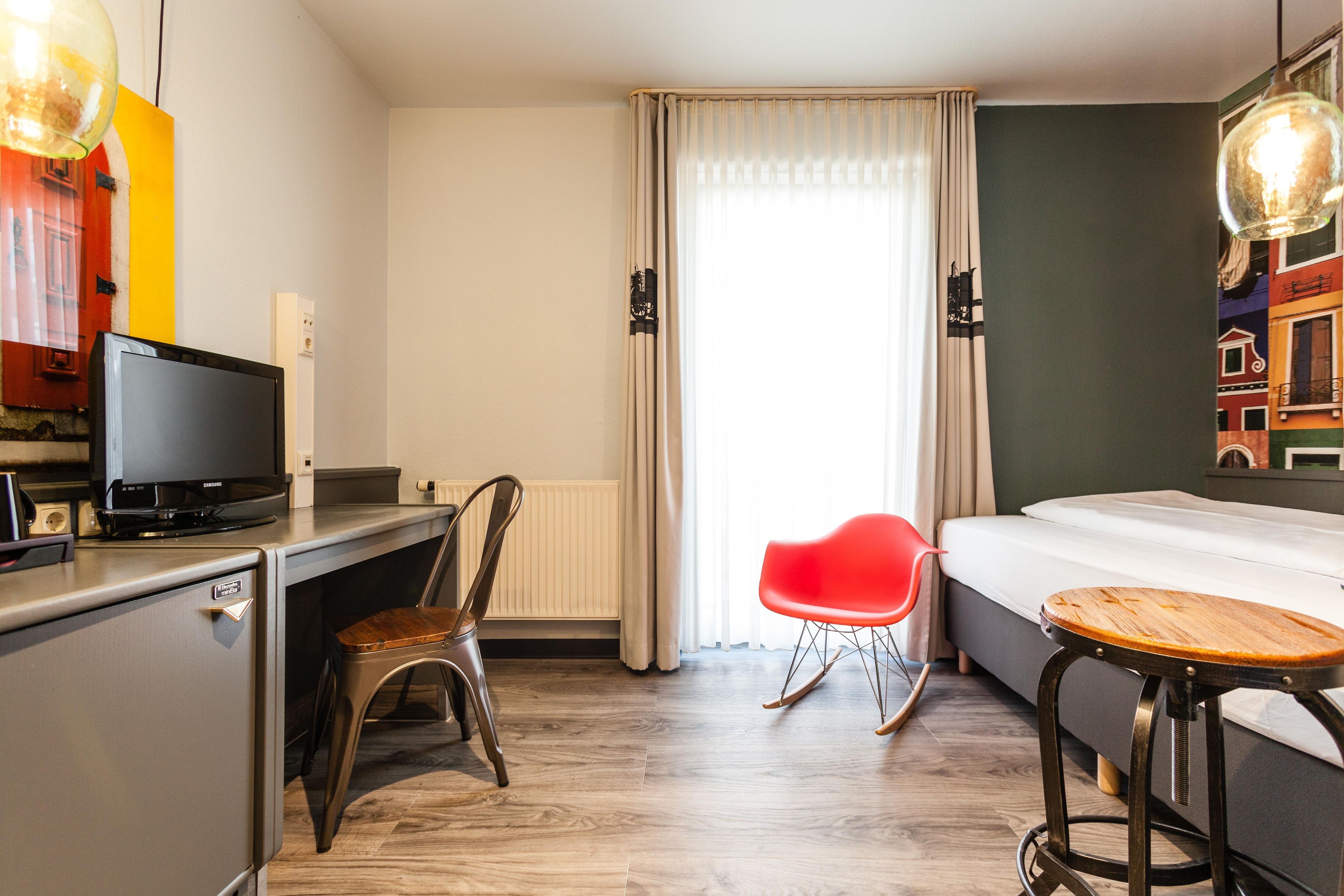 Una habitación de hotel con una cama, un escritorio con televisor, una silla roja y una mesita.