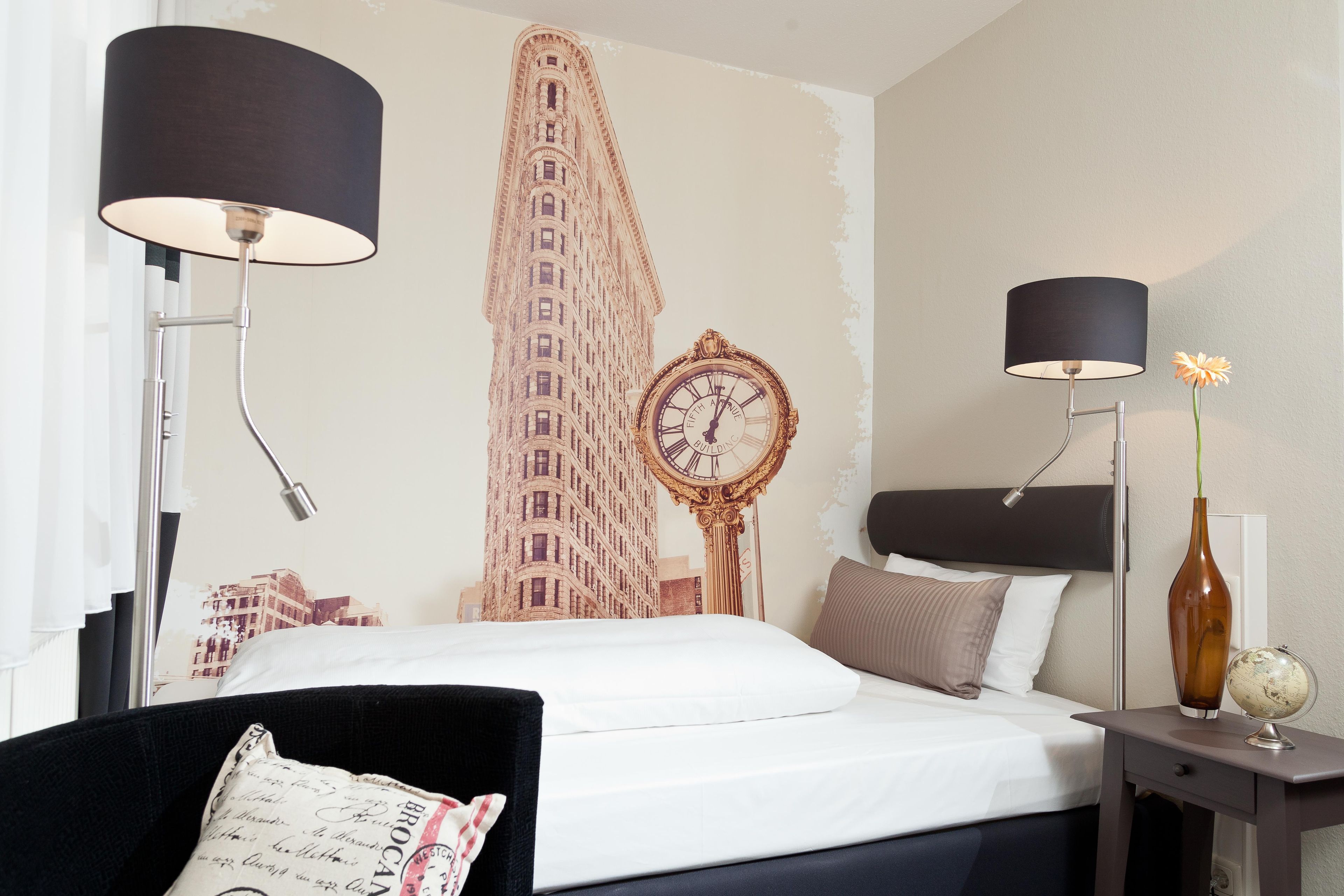 Ein Hotelzimmer mit einem Bett, zwei Nachttischlampen, einem Nachttisch mit Globus und einer Vase, einer Wanduhr und einem großen Wandgemälde eines Gebäudes.