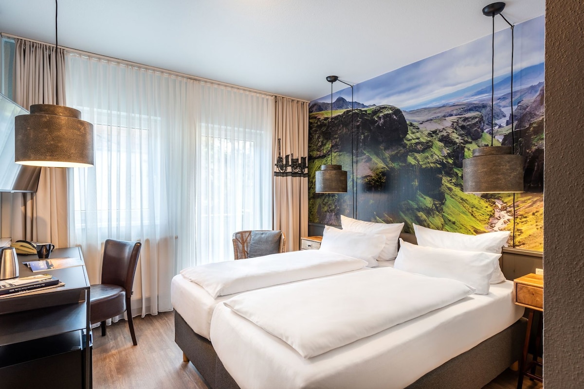 Habitación de hotel con una cama grande, un escritorio con silla, una lámpara y un mural decorativo en la pared.