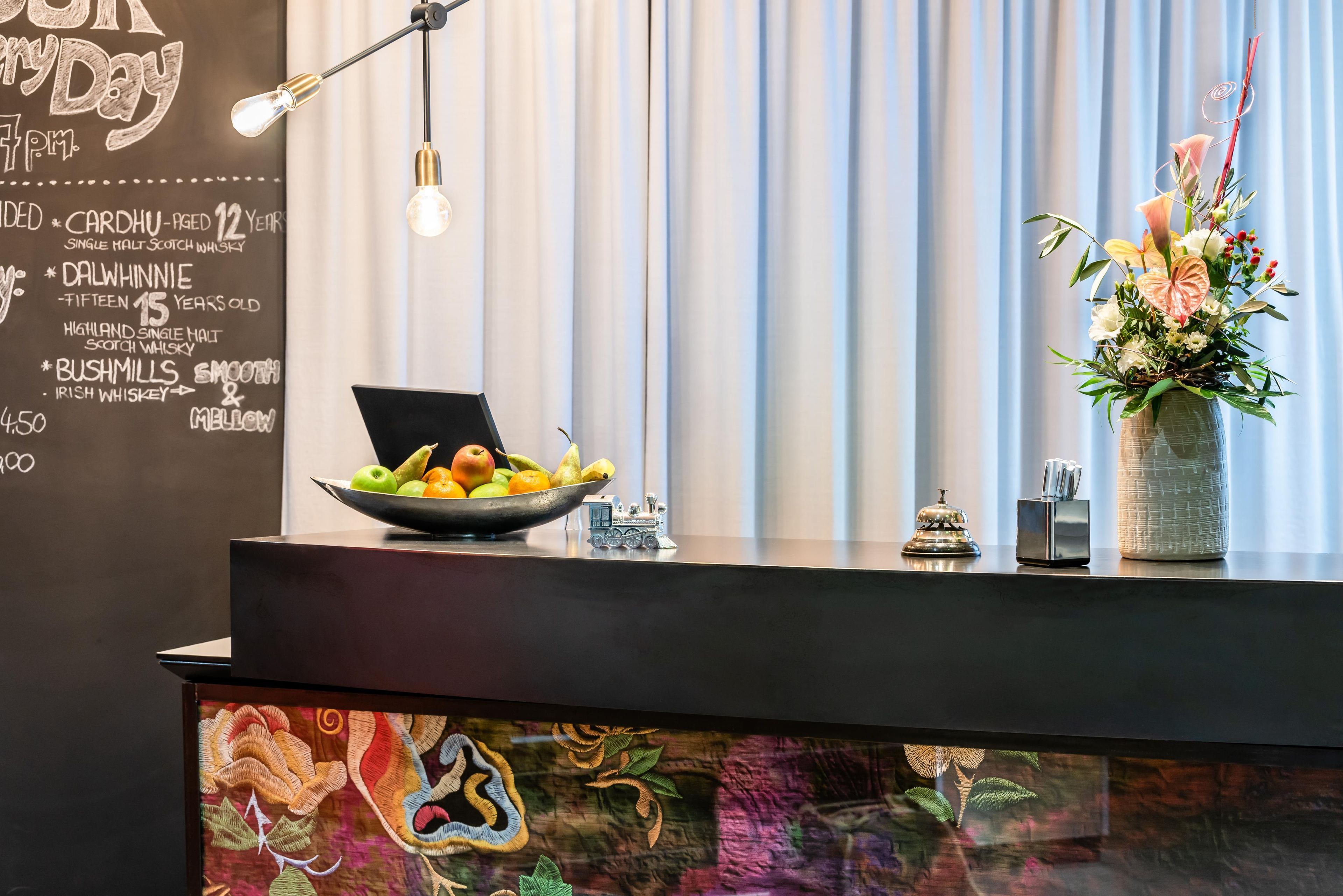 A reception desk with a chalkboard menu listing drinks, a bowl of fruit, a vase of flowers, and a decorative train model.