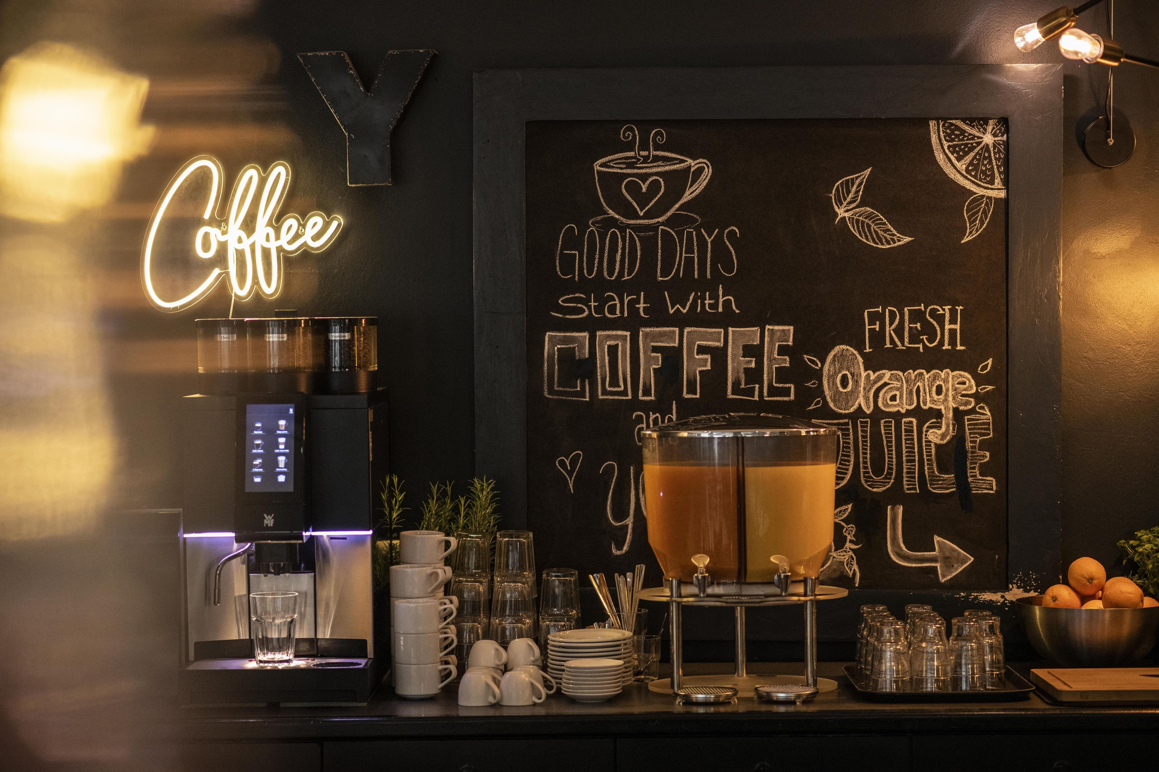 A coffee bar with a neon sign, a chalkboard with motivational writing, a coffee machine, and a large mug of coffee.