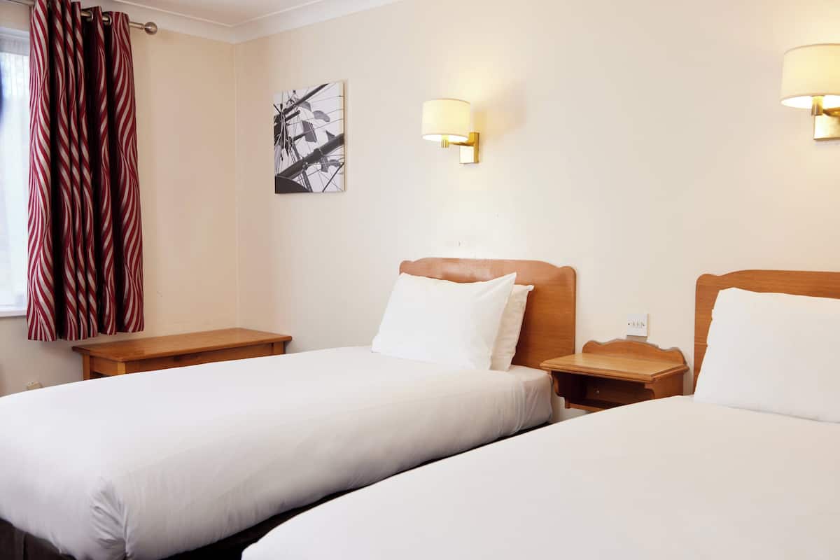 A hotel room with two single beds, wooden headboards, white bedding, a wooden nightstand, a lamp, and a wall-mounted artwork.