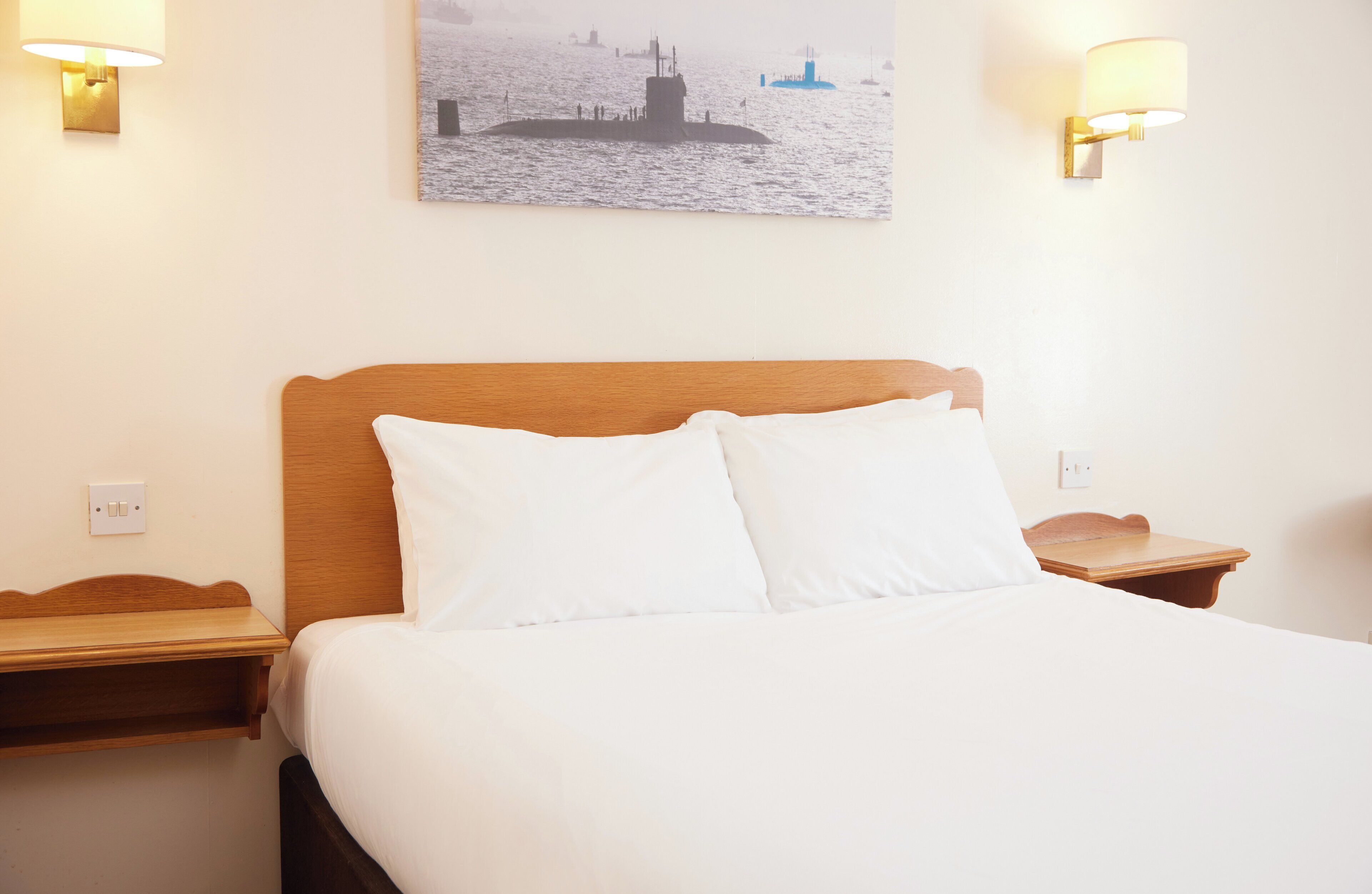 Una habitación de hotel con una cama, dos mesitas de noche, una lámpara de pared y un cuadro de un barco en la pared.