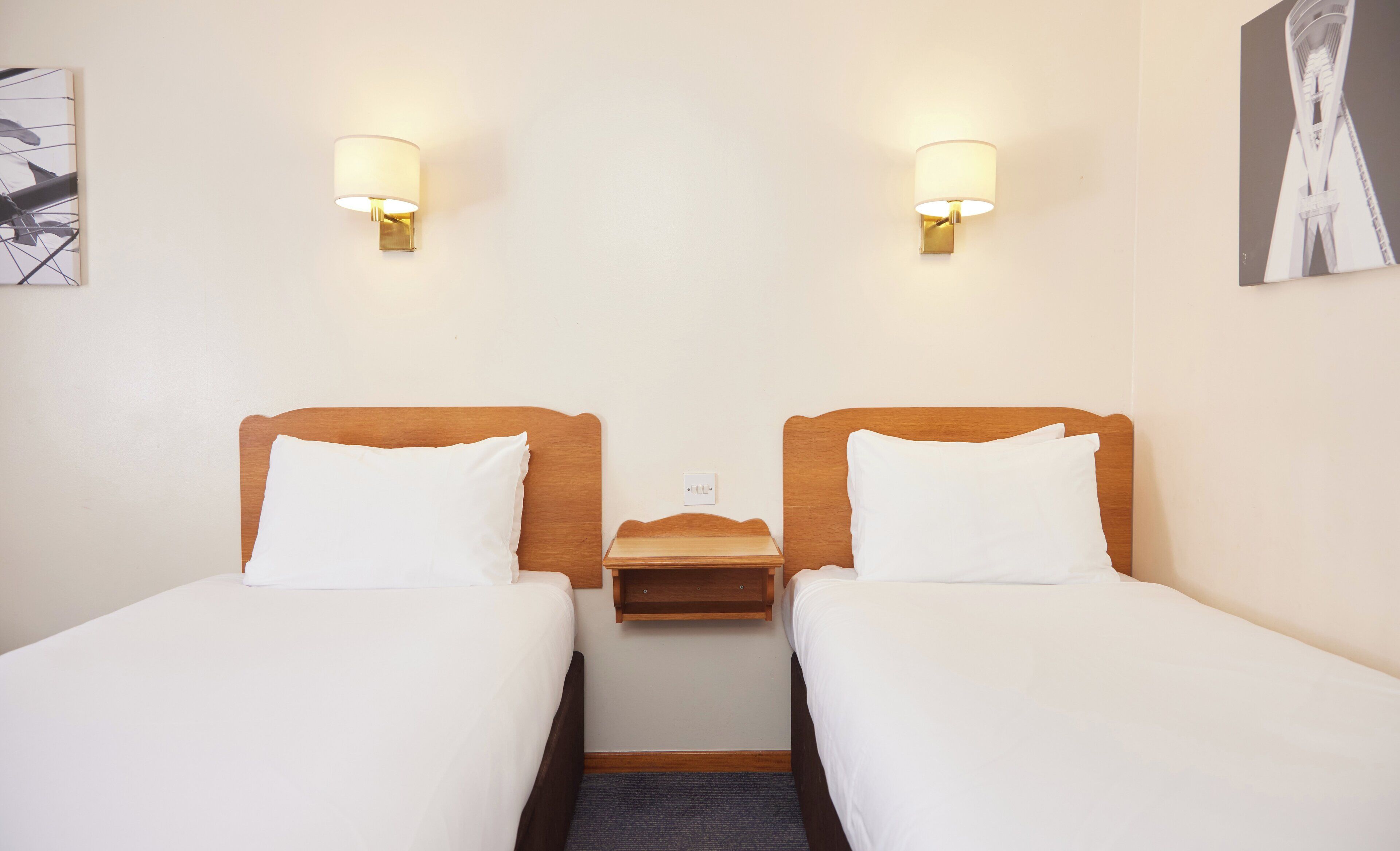 Habitación de hotel con dos camas individuales, una mesita de noche de madera y dos lámparas de pared.