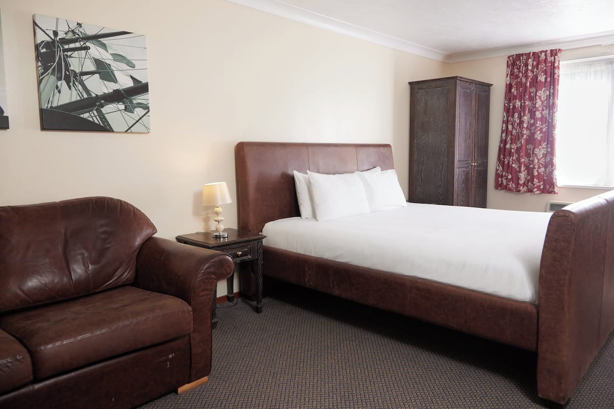 A hotel room with a large bed, a brown leather sofa, a nightstand with a lamp, and a wooden wardrobe.