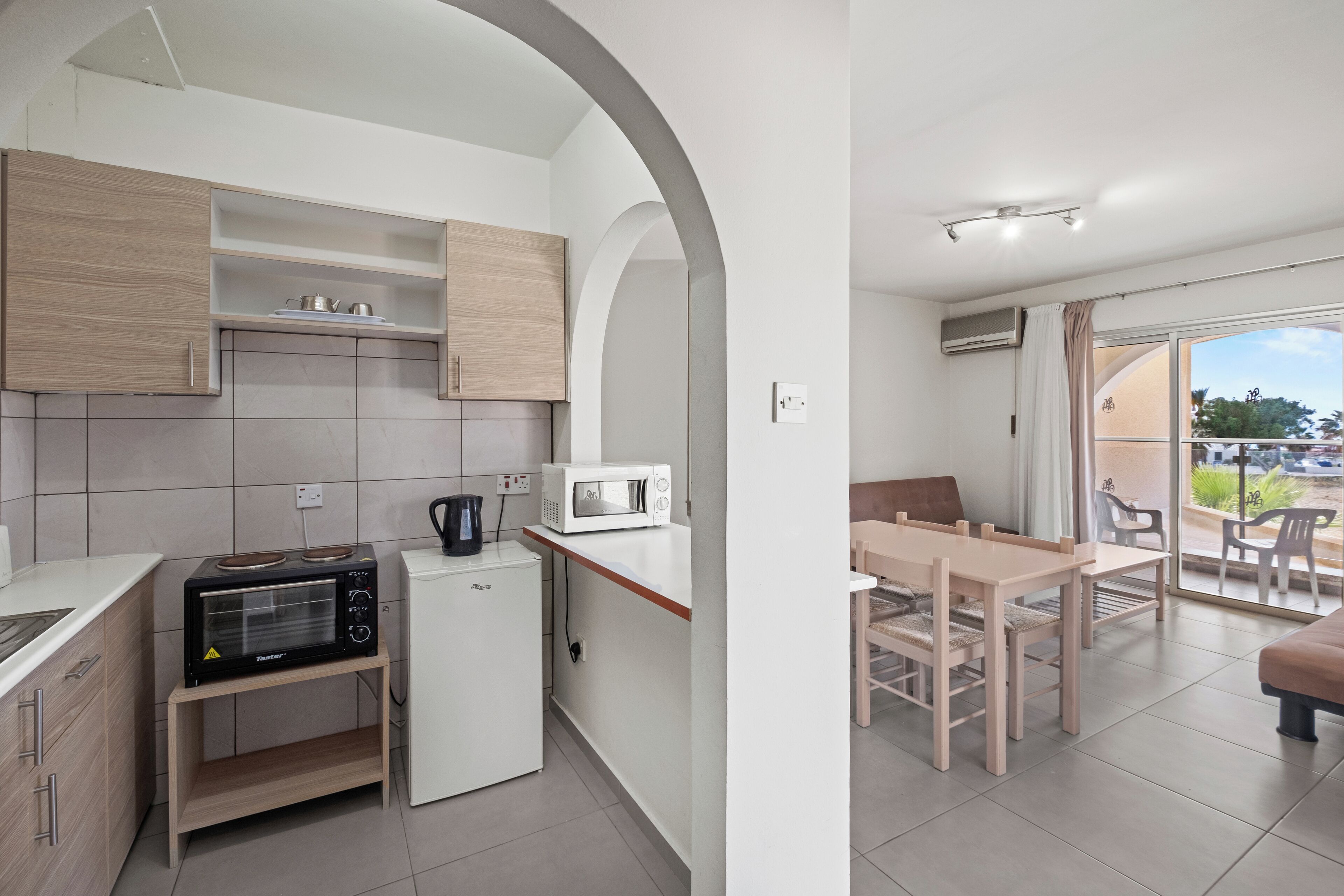A modern kitchen with a microwave, kettle, and a small refrigerator. There is a dining area with a table and chairs, and a balcony with a view.