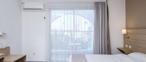 A hotel room with a bed, bedside tables, an air conditioner, a window with curtains, and a view of the outdoors.
