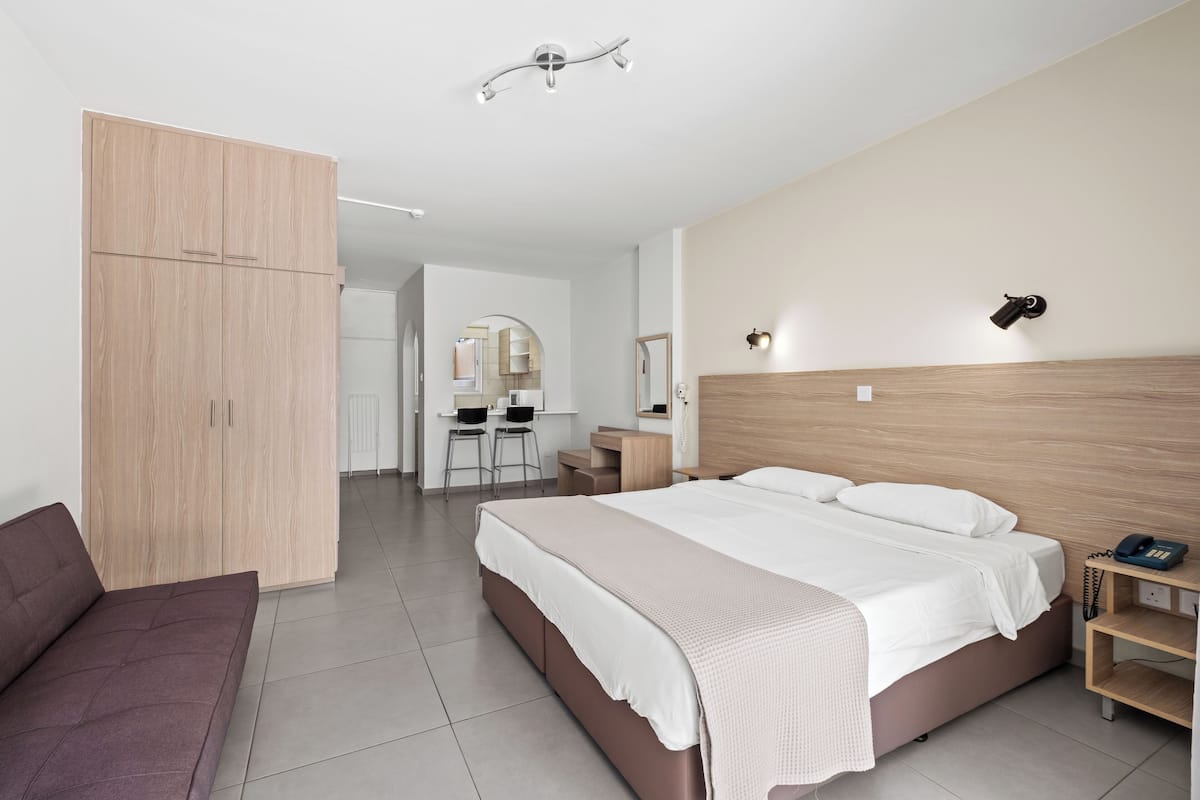 Quarto de hotel moderno com uma cama grande, um guarda-roupa de madeira, uma escrivaninha com duas cadeiras e uma pequena mesa de cabeceira com abajur.