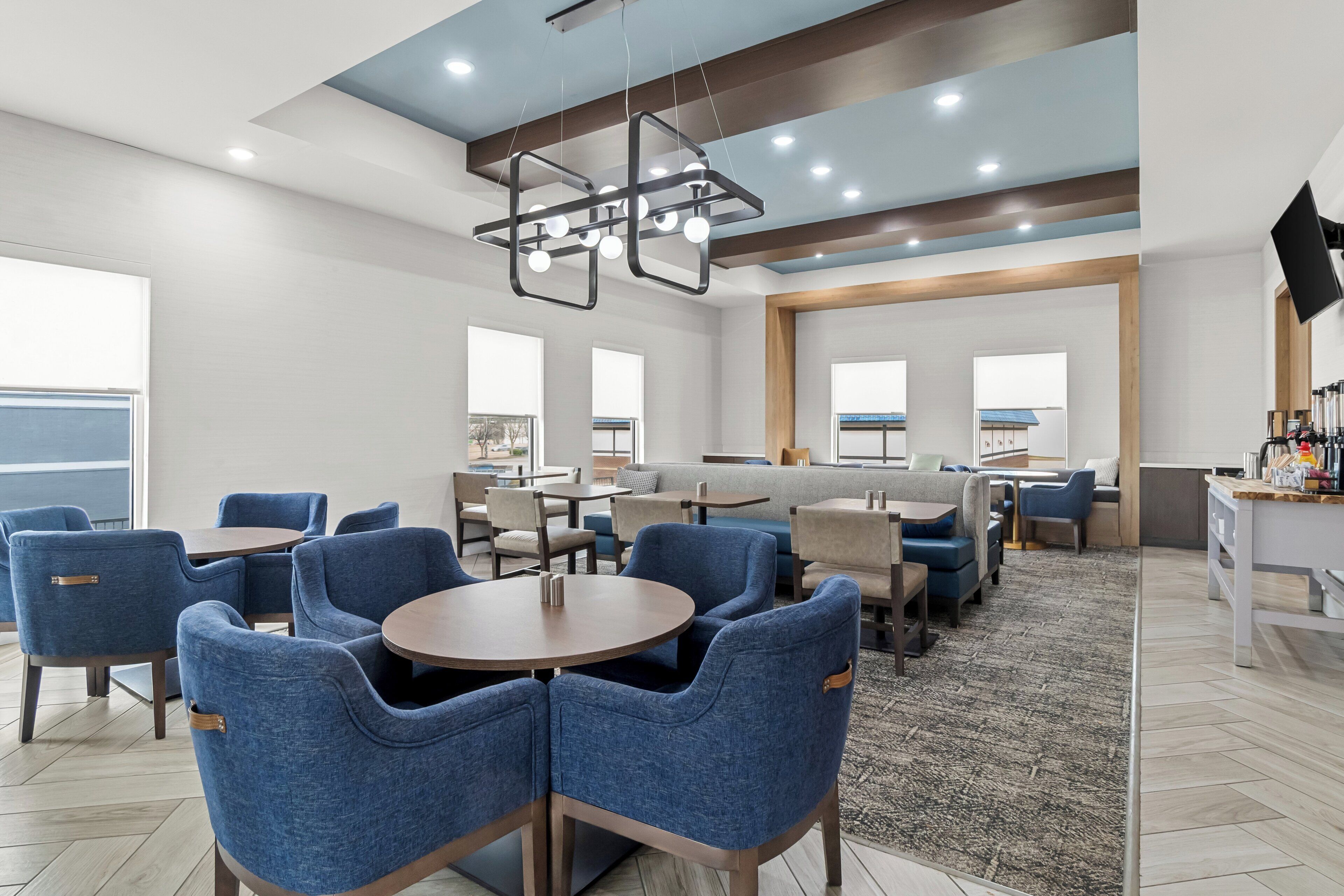 A modern hotel lobby with round tables, blue upholstered chairs, a large seating area, and a flat-screen TV mounted on the wall.