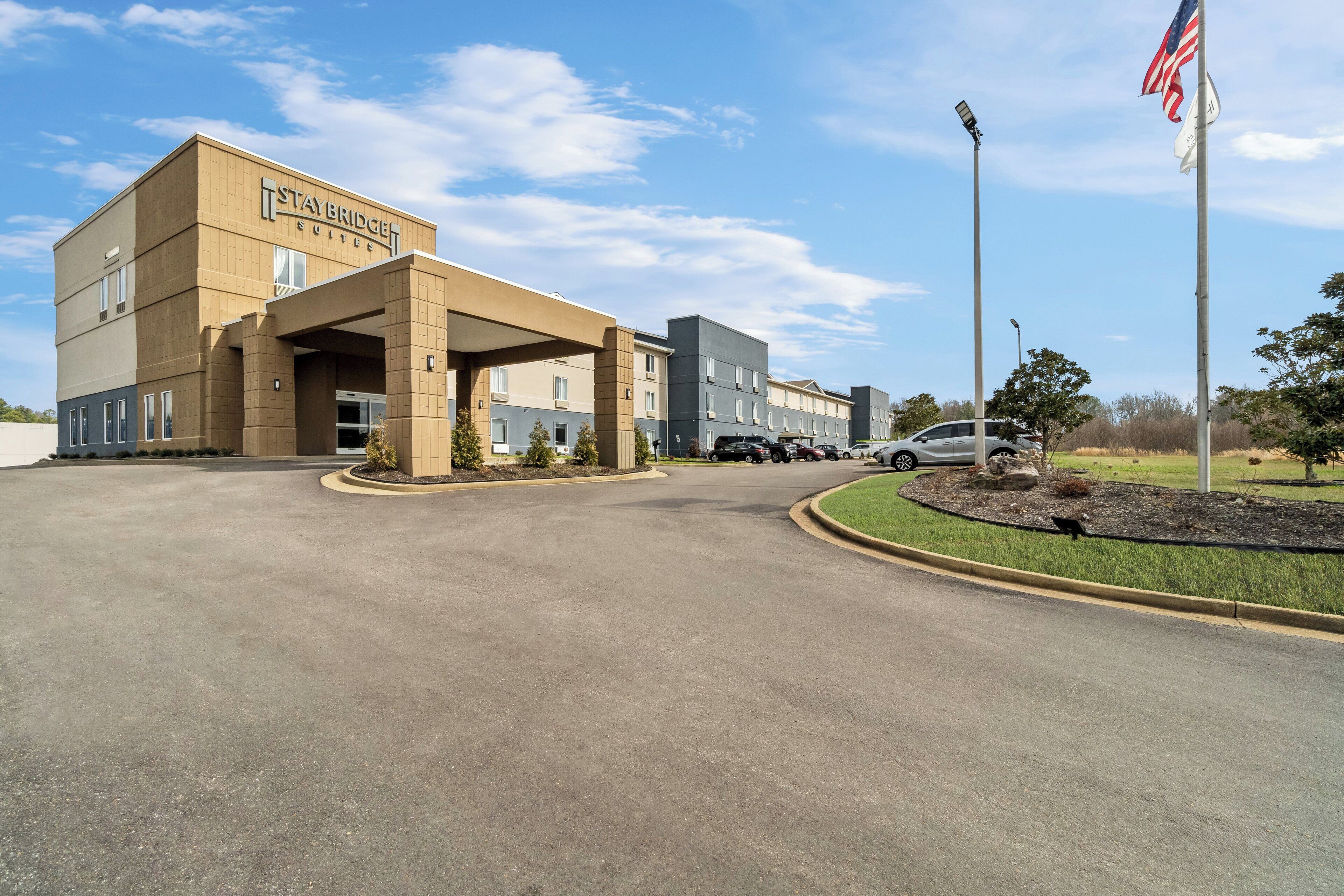 A Staybridge Suites hotel with a covered entrance, a parking lot, and an American flag.