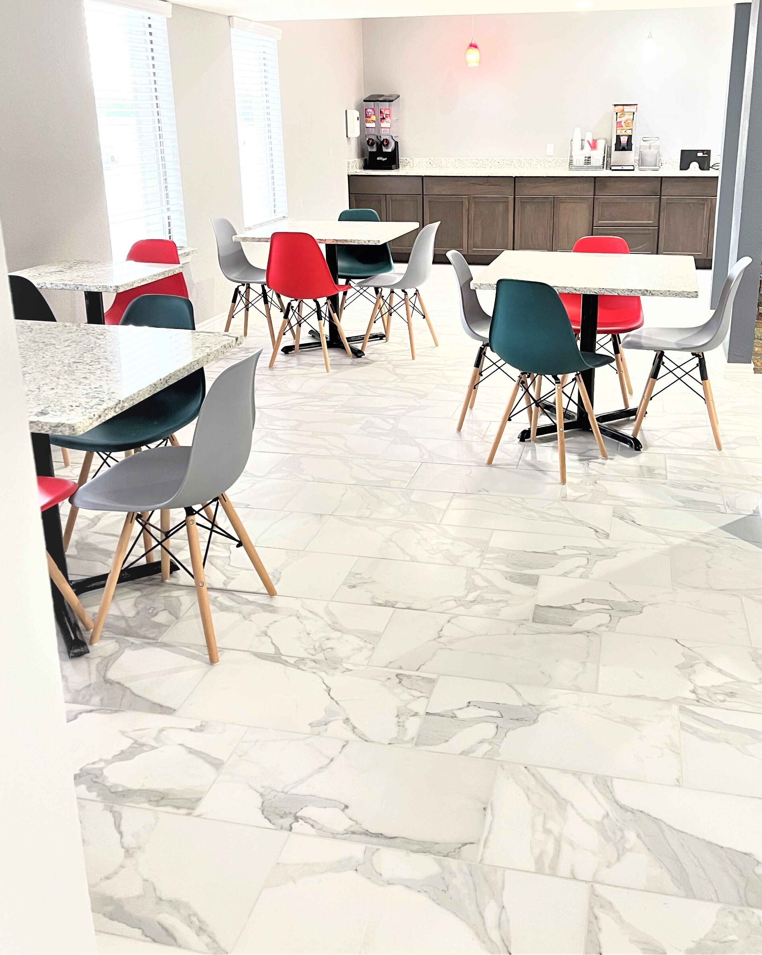 Un comedor moderno con suelo de mármol, sillas rojas y grises, y una cocina al fondo.