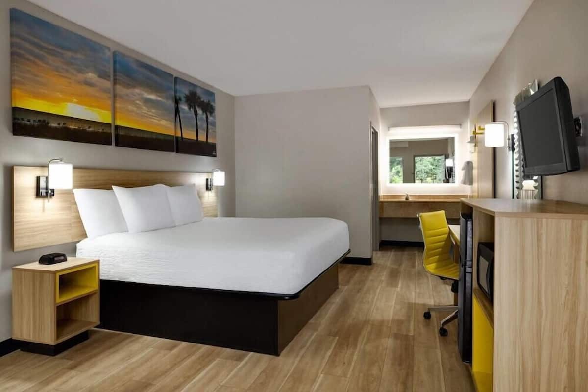 A modern hotel room with a bed, bedside tables, a desk, a chair, a TV, and a painting on the wall.