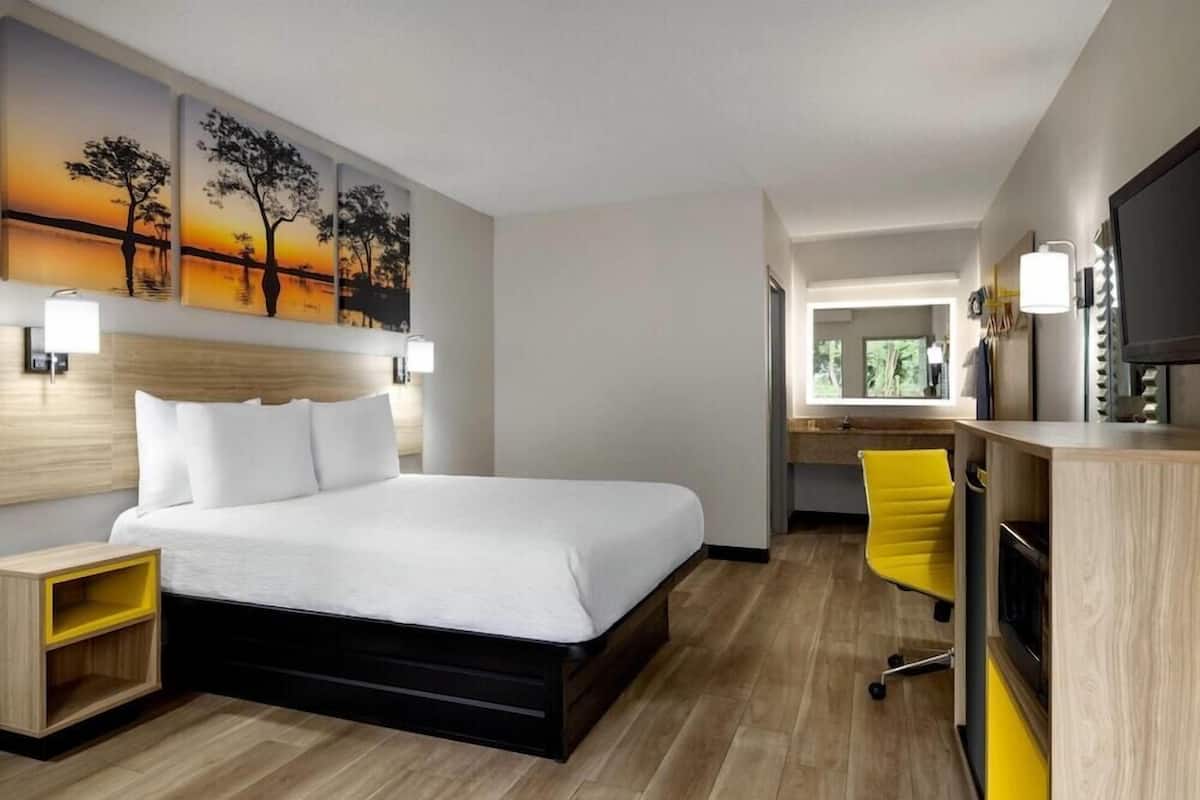 A hotel room with a bed, bedside table, desk, chair, and artwork on the wall.