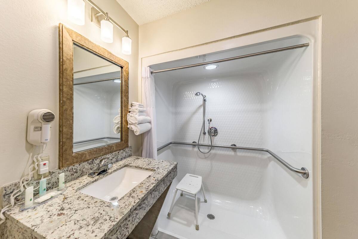 Une salle de bain équipée d’un lavabo, d’un miroir, d’un porte-serviettes, d’une douche avec des barres d’appui et d’un petit marchepied.