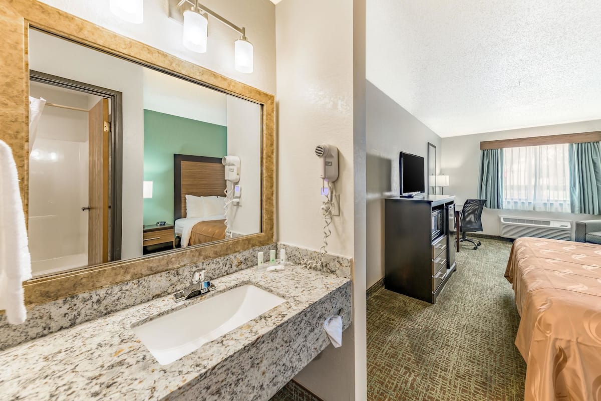Une salle de bain d’hôtel équipée d’un lavabo, d’un miroir et d’un sèche-cheveux. La chambre comprend un lit, un bureau et une télévision.