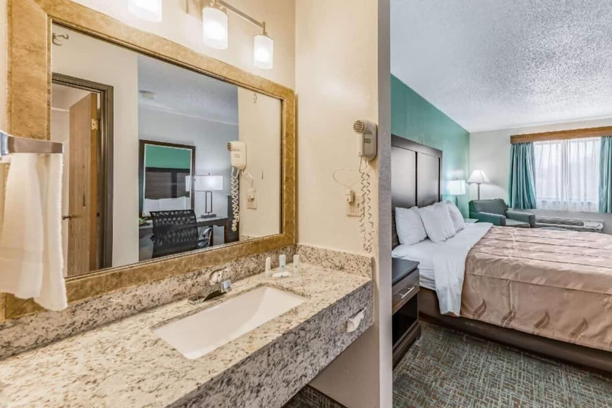 Une chambre d’hôtel avec un meuble-lavabo en marbre dans la salle de bain, un grand lit, une table de chevet et une vue sur la pièce depuis la fenêtre.
