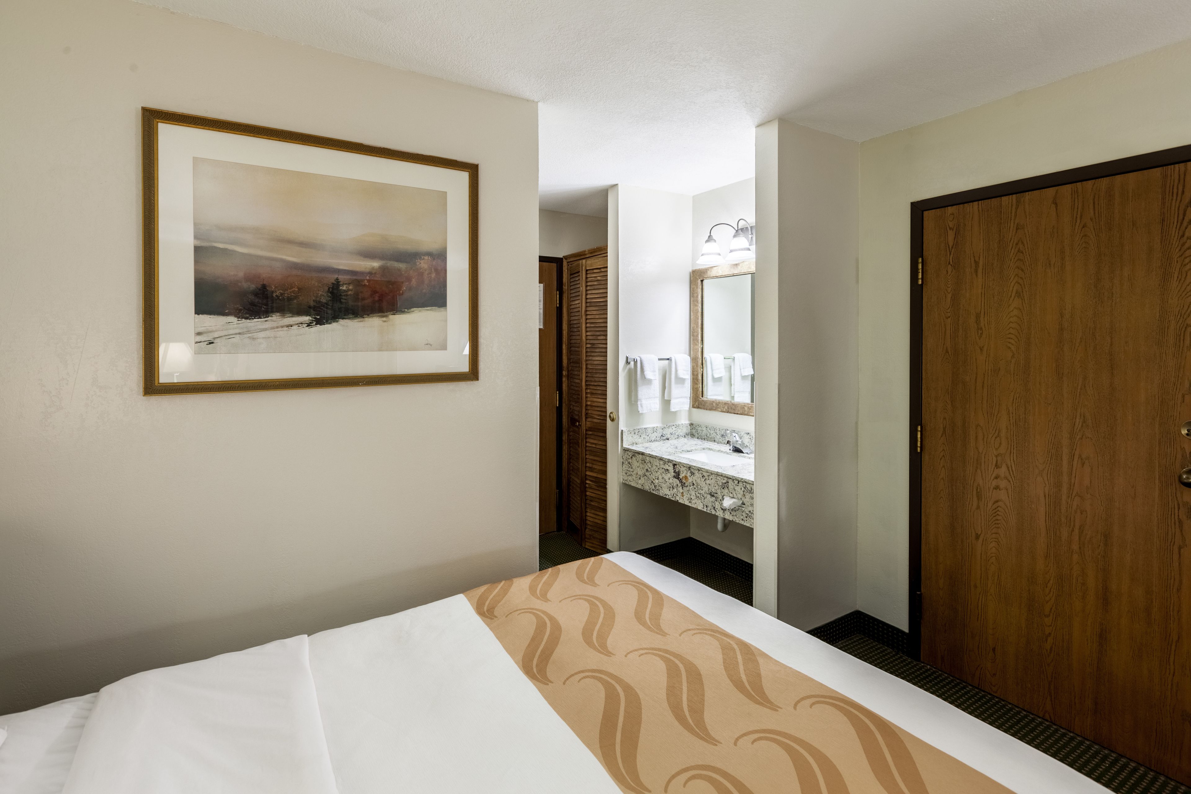 A hotel room with a bed, a bathroom with a sink and mirror, and a wooden door.