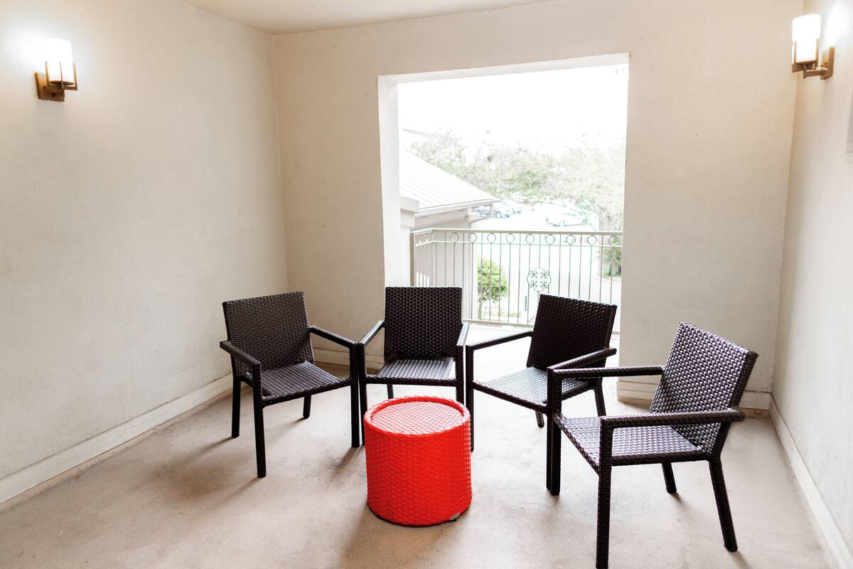 Quarto nos fundos com quatro cadeiras de vime, um objeto cilíndrico vermelho e uma janela com vista para uma casa.