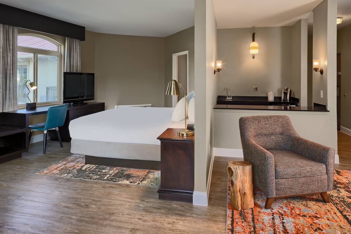 Quarto de hotel moderno com uma cama grande, uma escrivaninha com televisão, uma cadeira, uma pequena copa e um banheiro com espelho e iluminação.