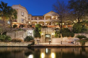 Un hôtel à plusieurs étages avec une piscine en premier plan, des murs en pierre et diverses plantes.