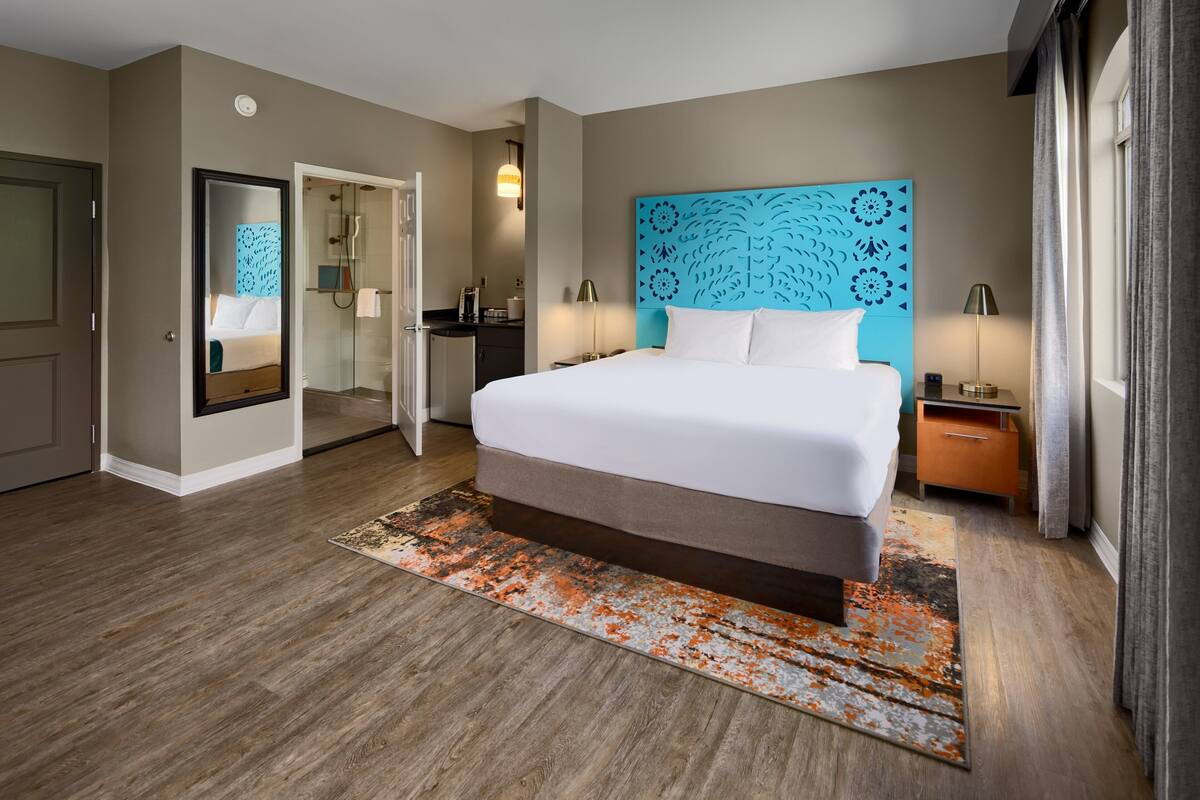 Quarto de hotel com uma cama grande, um espelho, banheiro com chuveiro e mesa de cabeceira com abajur.