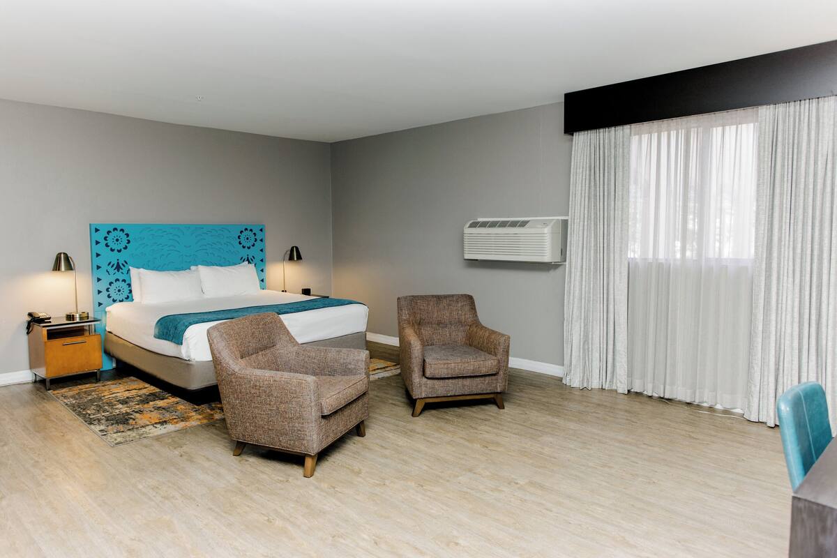 Quarto de hotel com uma cama, duas poltronas, uma mesa de cabeceira, um abajur, um ar condicionado e uma janela com cortinas.