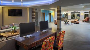 Eine moderne Hotelhalle mit einem Holzbüro, zwei Monitoren, bunten Stühlen, einer Couch und einem an der Wand montierten Flachbildfernseher.
