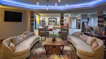 A modern living room with sofas, a coffee table, and a TV. There is a dining area in the background with a high ceiling and pendant lighting.