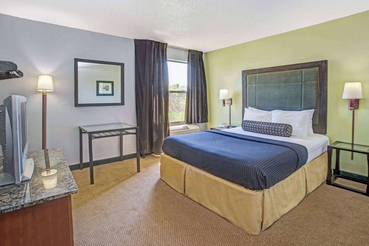 A hotel room with a bed, bedside lamps, a small table, a mirror, and a window with curtains.