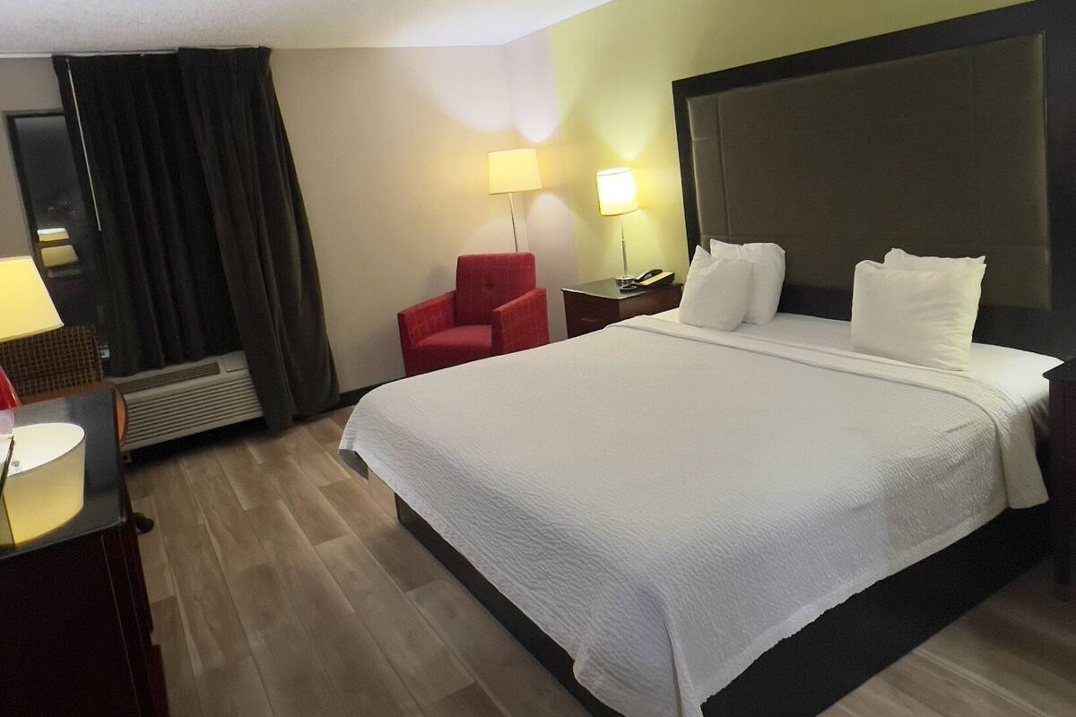 A hotel room with a large bed, two bedside lamps, a red chair, a desk with a lamp, and a mirror.