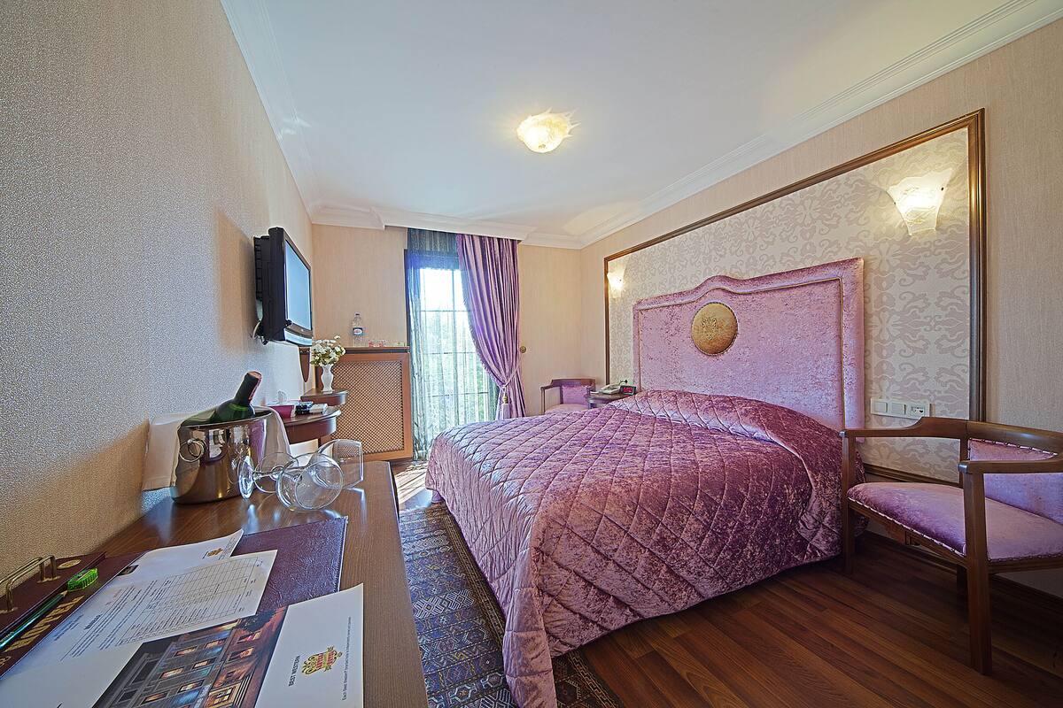 A hotel room with a large bed, a wooden desk, a television mounted on the wall, and a window with curtains.