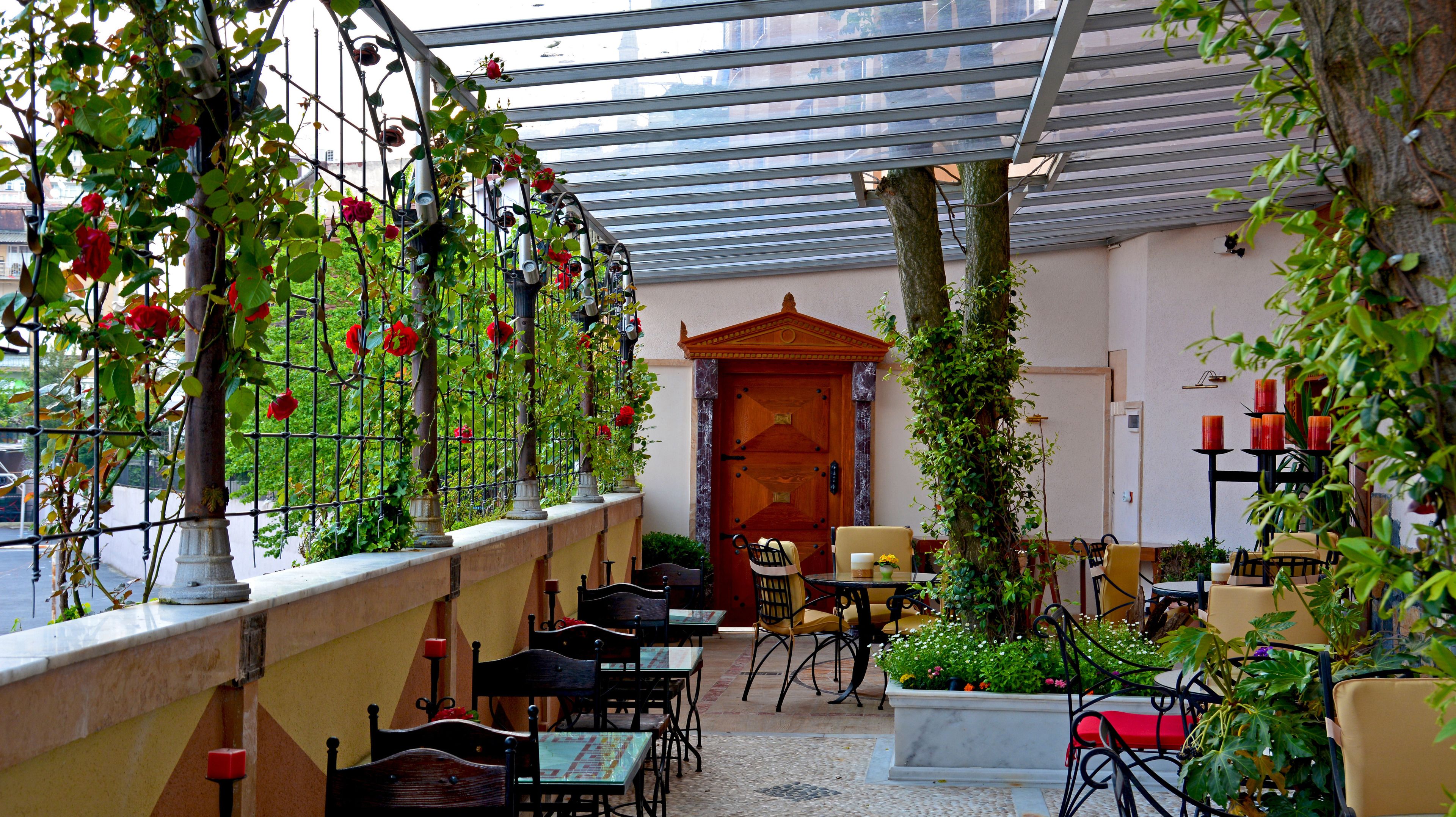 A covered outdoor seating area with tables and chairs, surrounded by greenery and flowers.