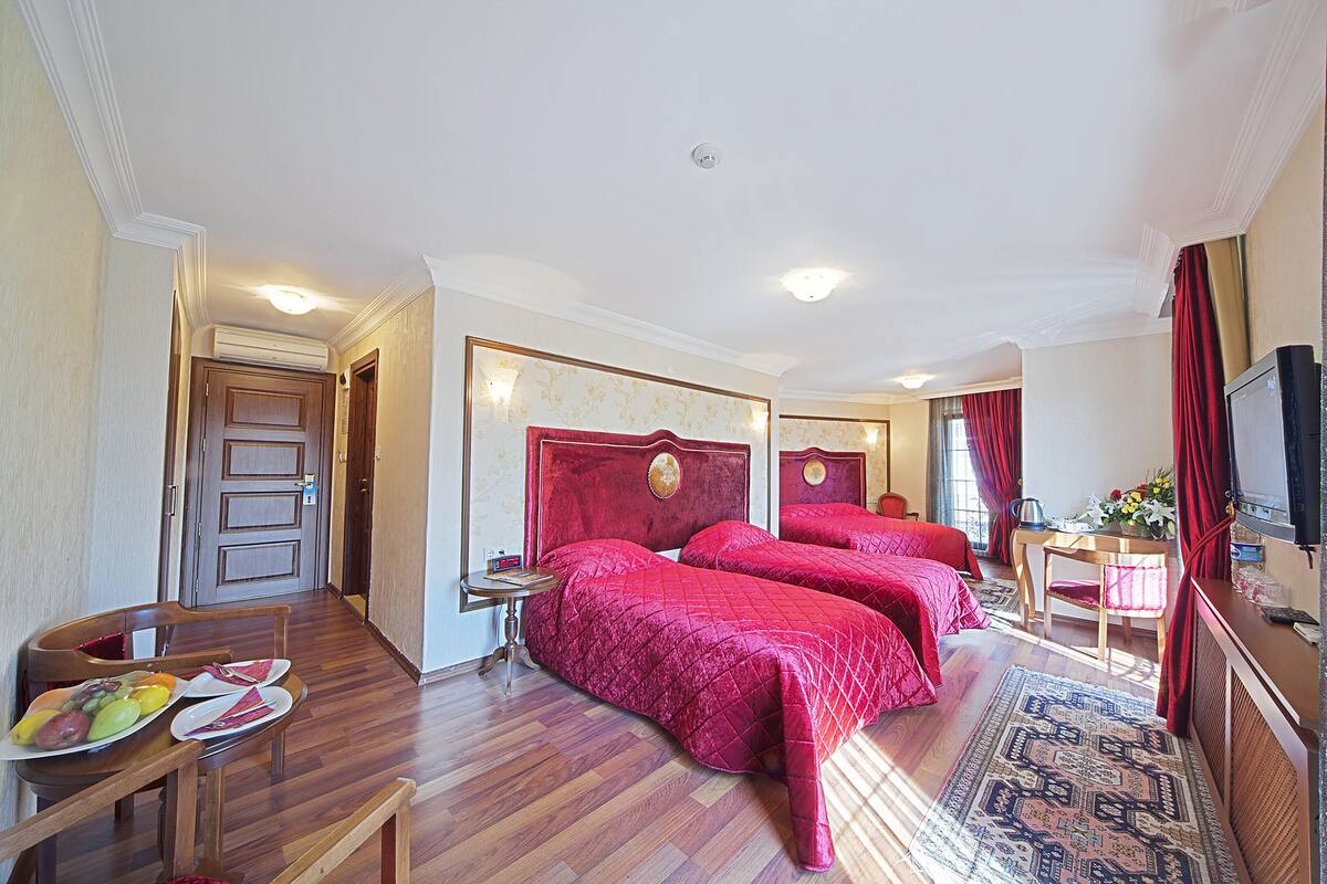 A hotel room with two beds, a dining table with plates of fruit, a small table with a vase, and a television on the right.