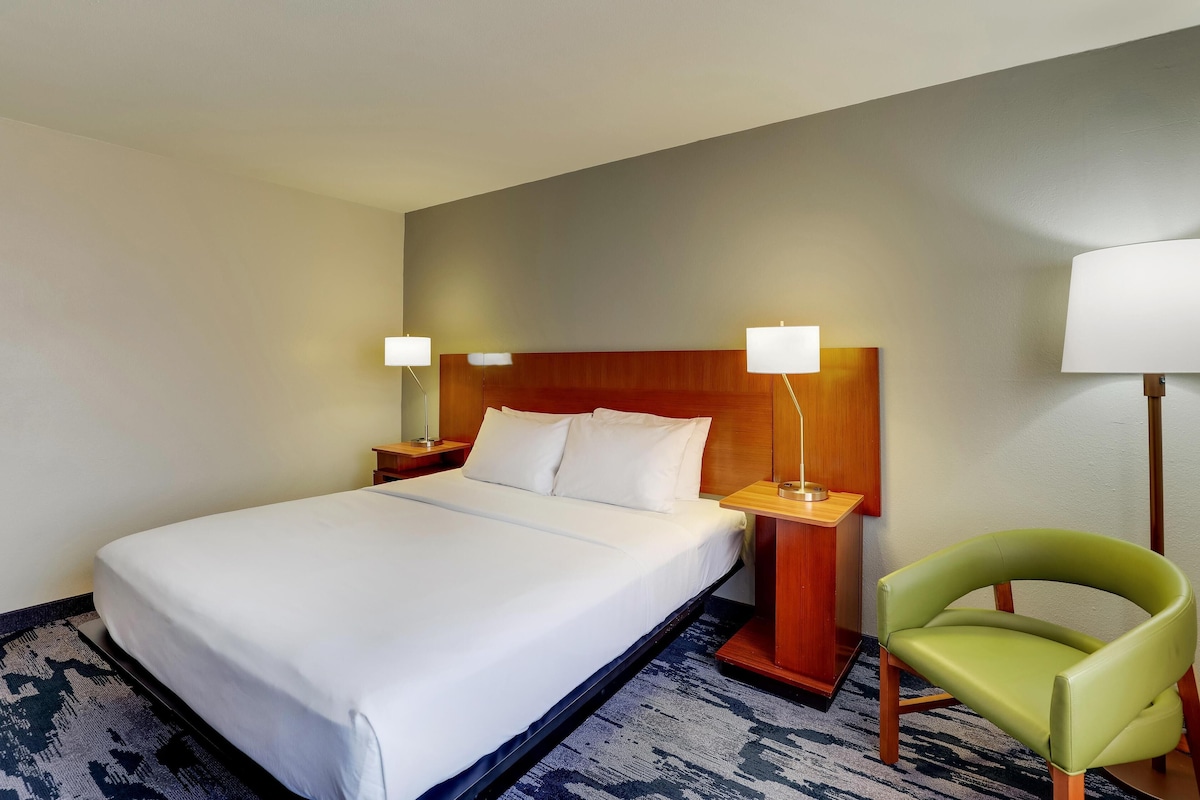 A hotel room with a large bed, two bedside lamps, a green chair, and a wooden headboard.