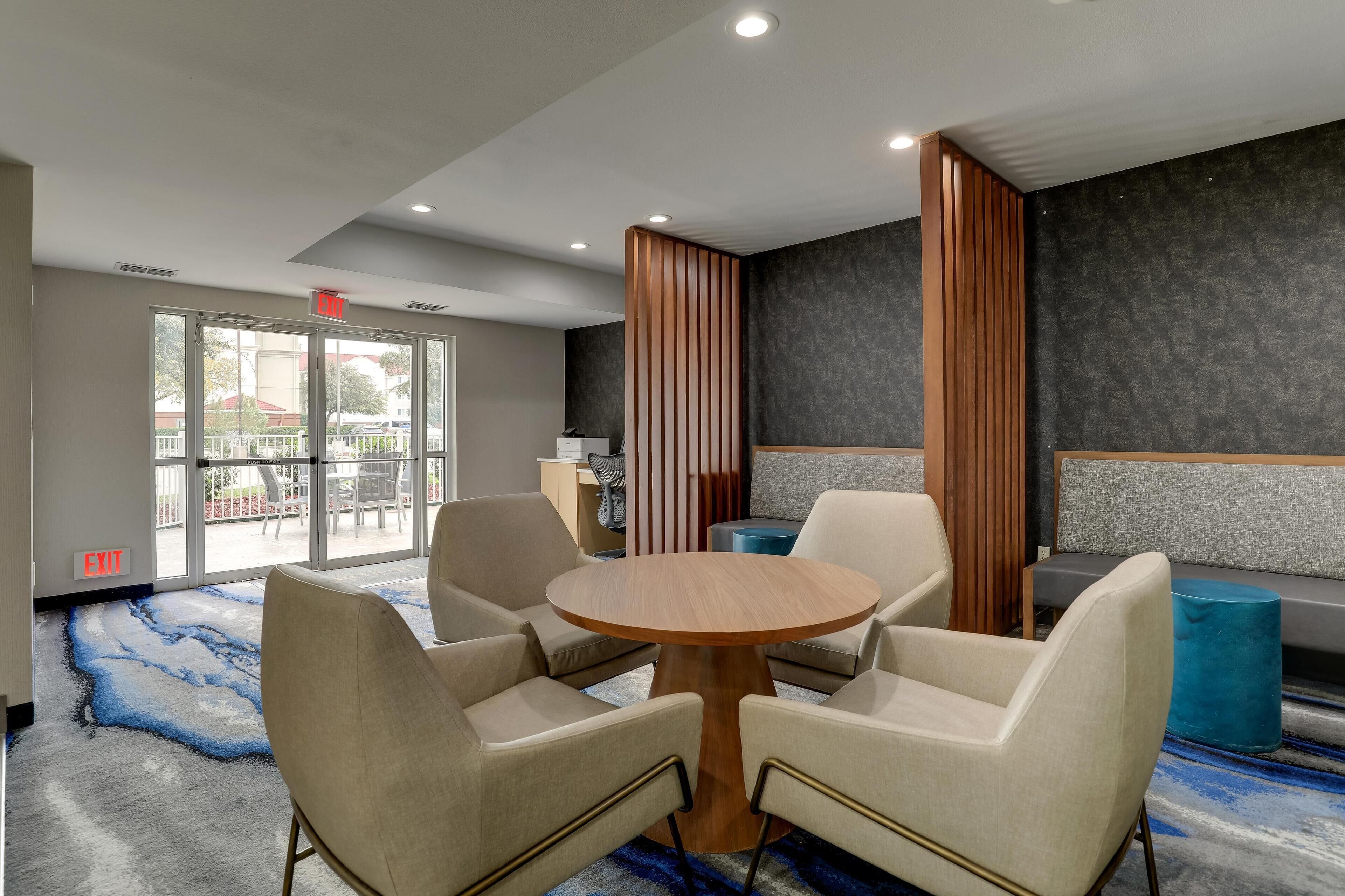 Una moderna hall d'hotel con un tavolo rotondo in legno circondato da quattro poltrone beige, un divano grigio e un poggiapiedi blu. Si vede una reception con un computer e una sedia, e un'ampia finestra con un'indicazione di uscita.