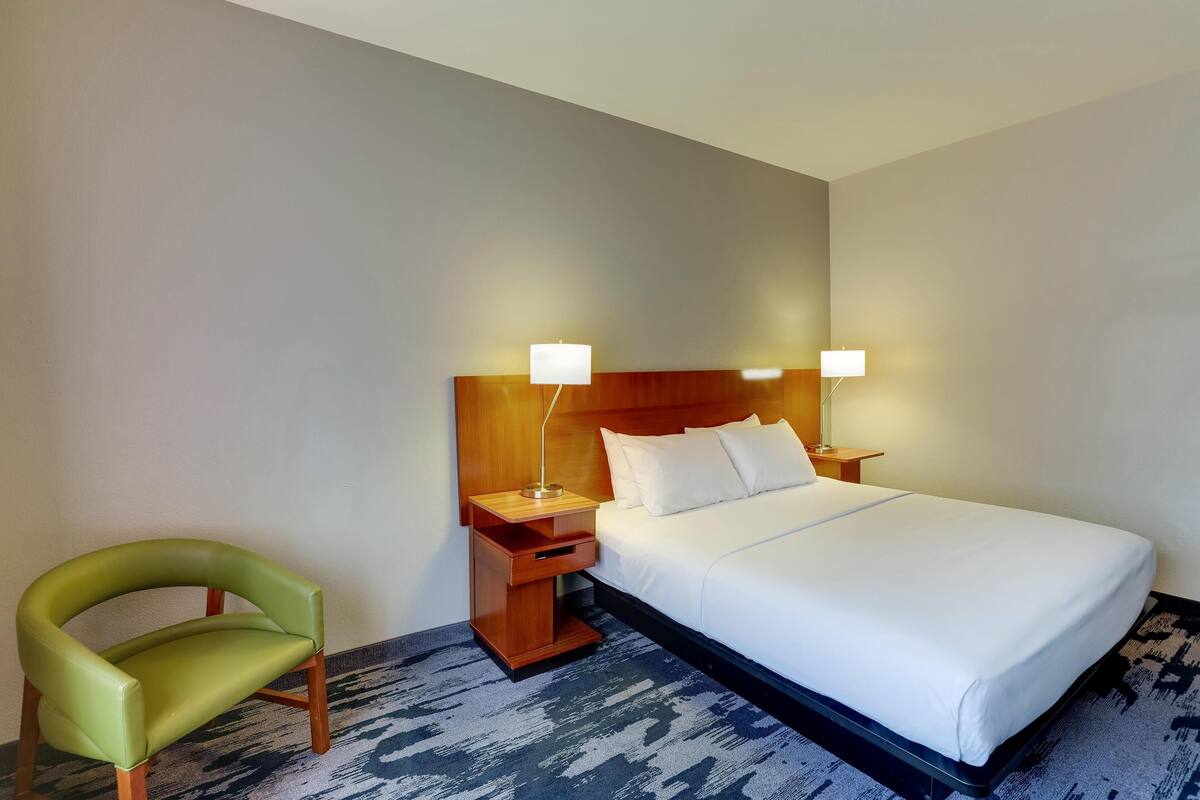 A hotel room with a bed, a green chair, two bedside tables with lamps, and a patterned carpet.
