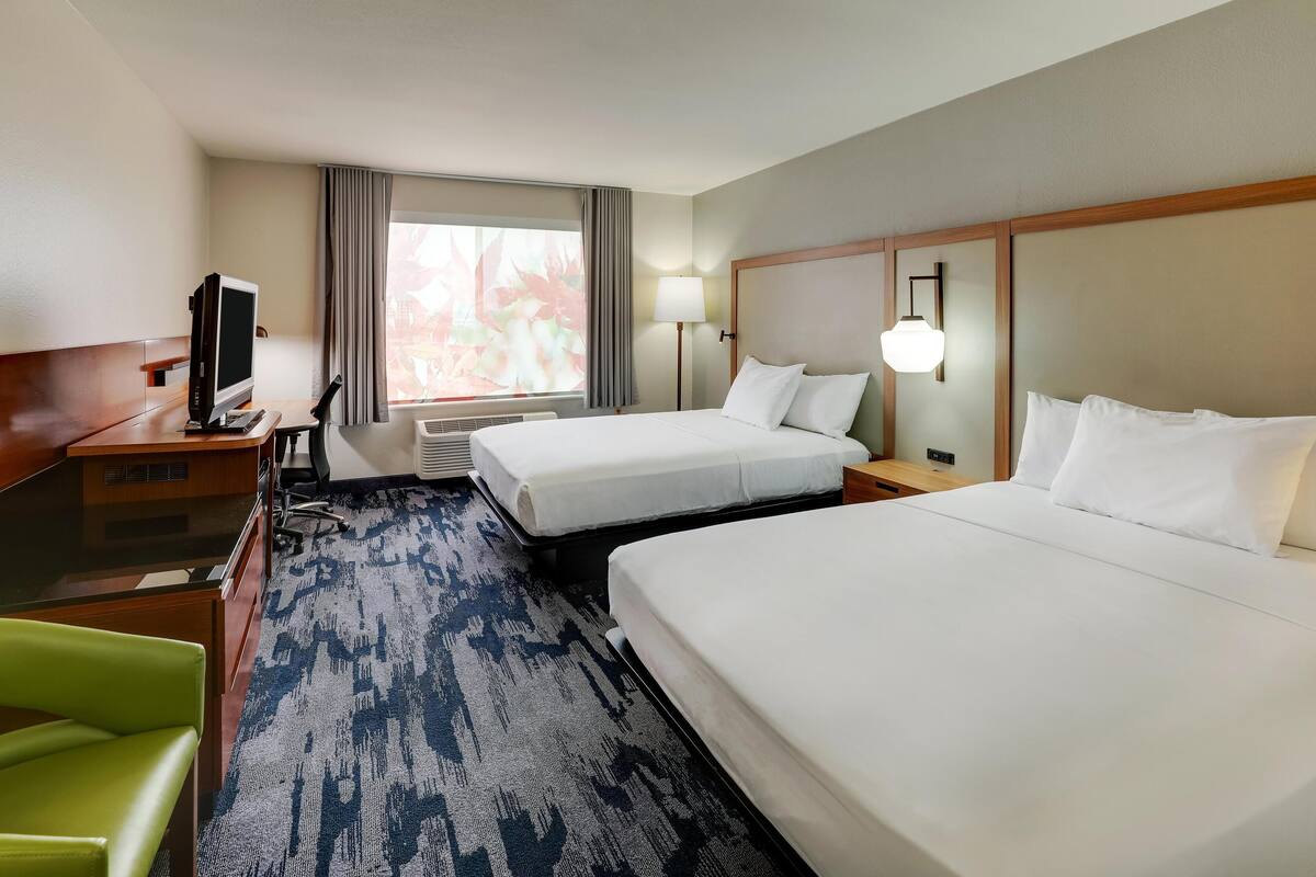 A hotel room with two beds, a desk with a computer, a green chair, and a window with curtains.