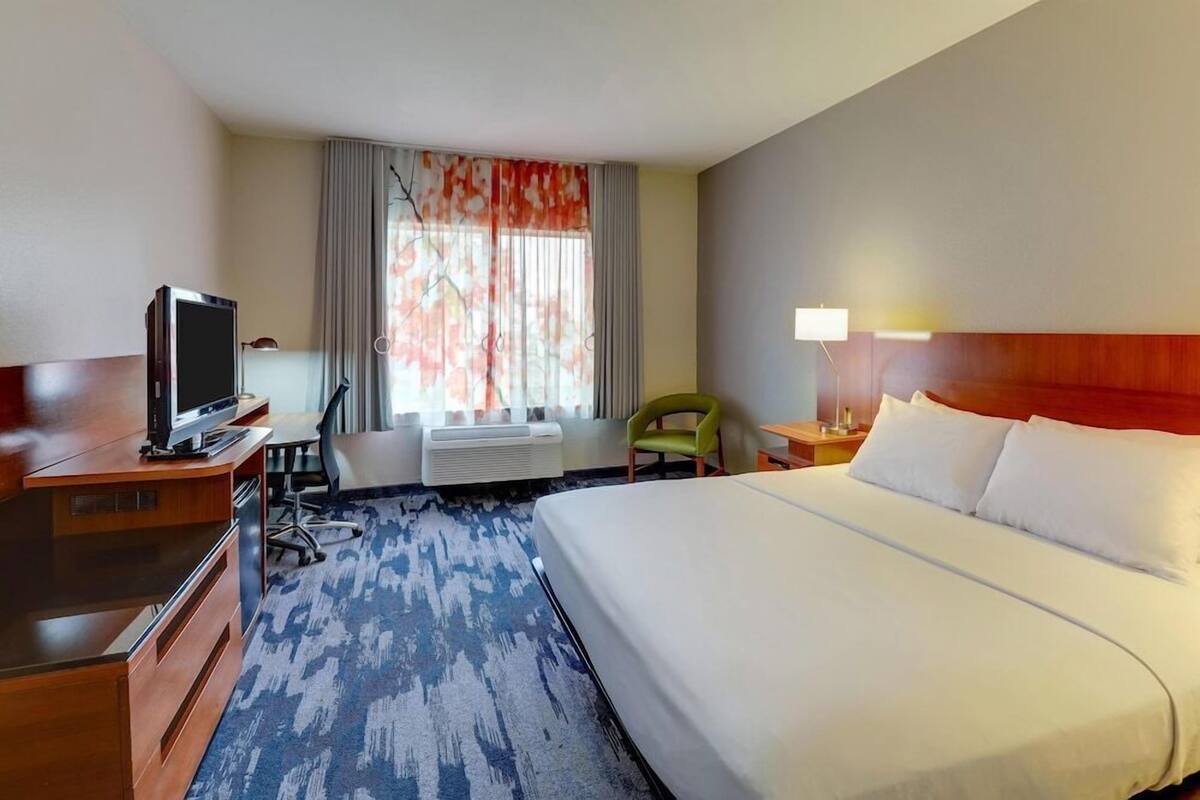 A hotel room with a bed, a desk with a television, a chair, a lamp, and a window with curtains.