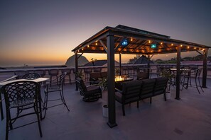 An outdoor seating area with tables and chairs, a covered gazebo, and a view of the ocean at dusk. - Ascot Suites (Morro Bay)