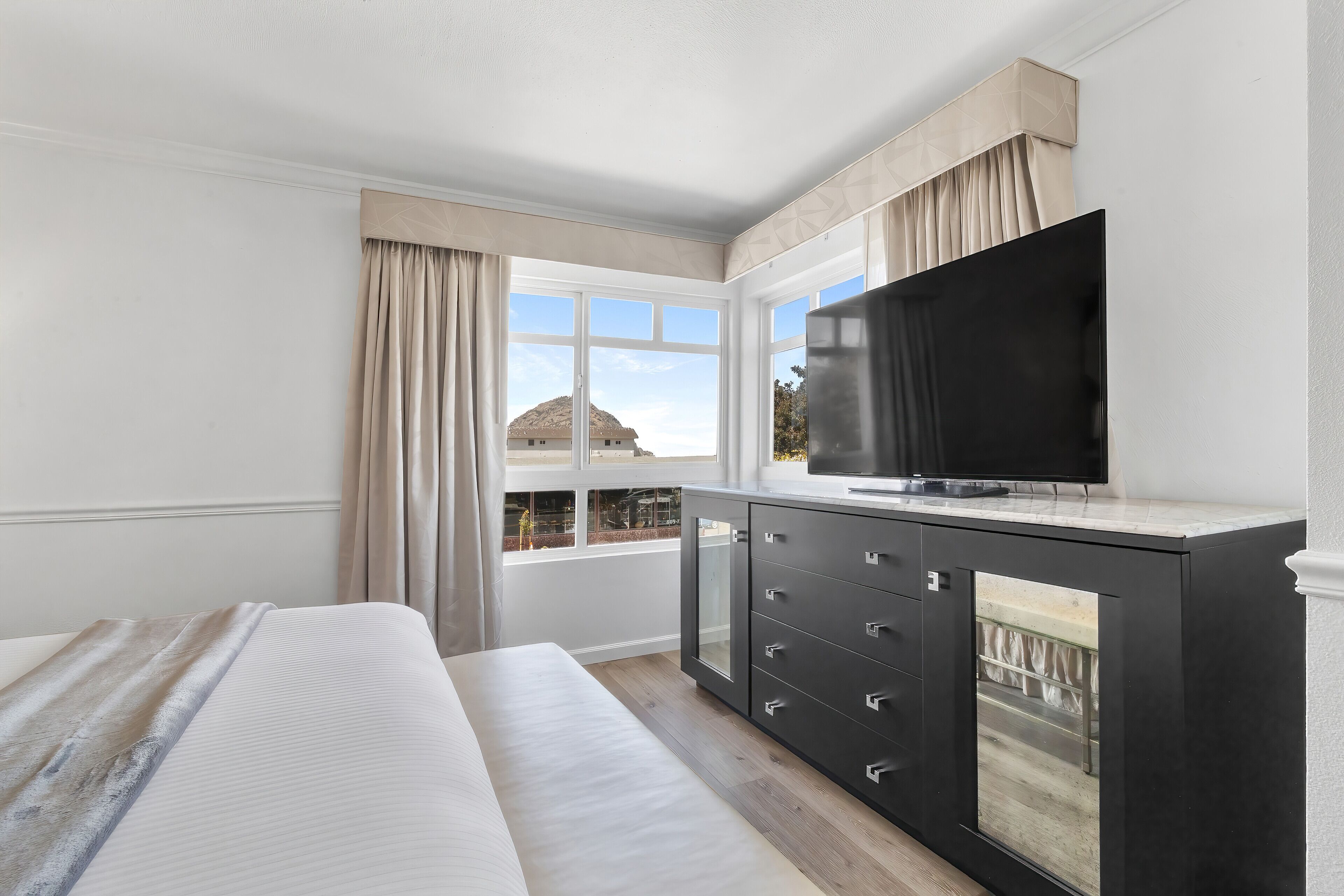 Un dormitorio con cama, televisor sobre un mueble negro y vista a un edificio a través de ventanas amplias.