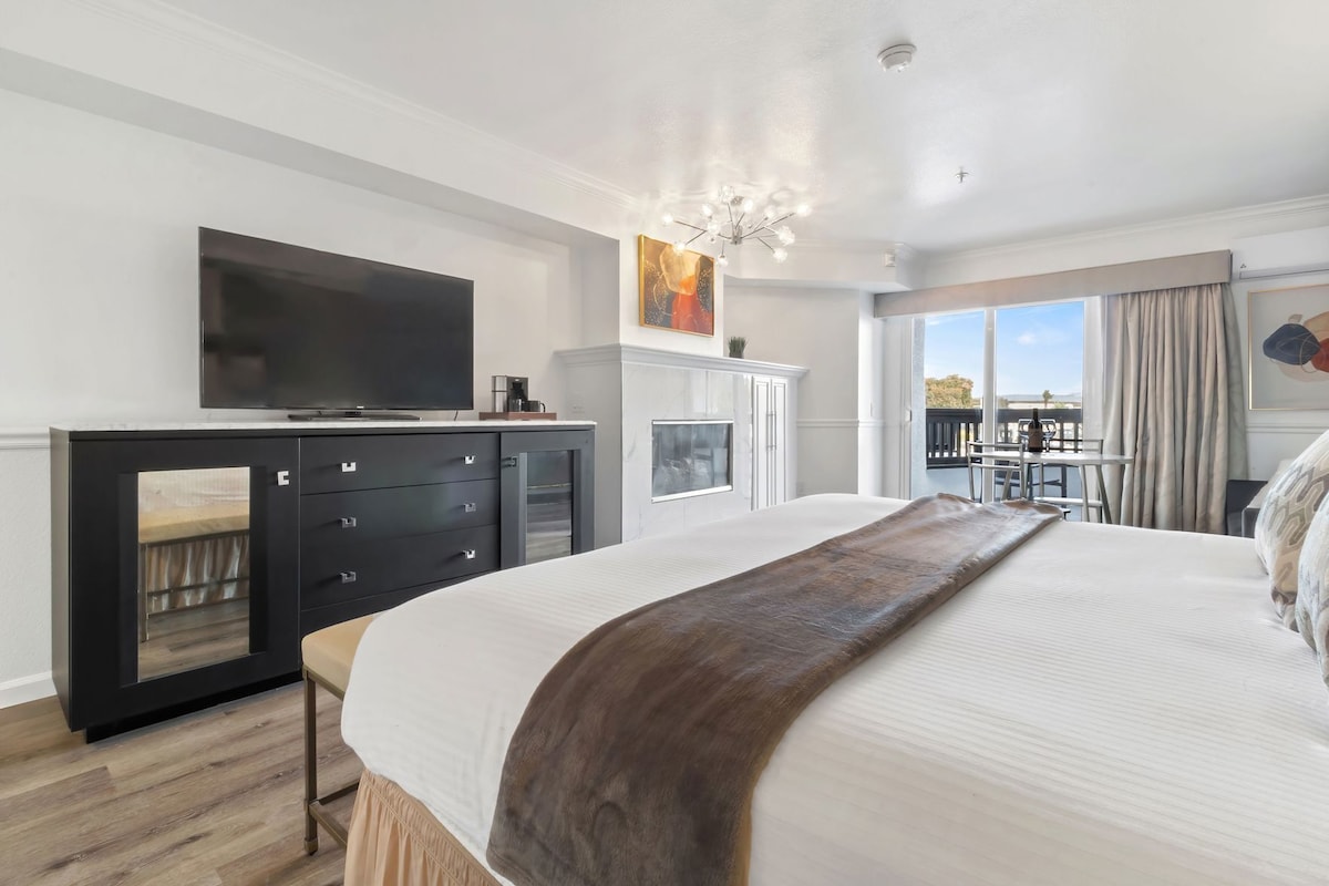 A modern hotel room with a large bed, a TV, a minibar, and a balcony with a view.