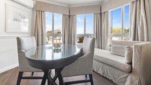 Habitación de esquina con mesa de vidrio, dos sillas, un sofá y ventanas amplias con cortinas.