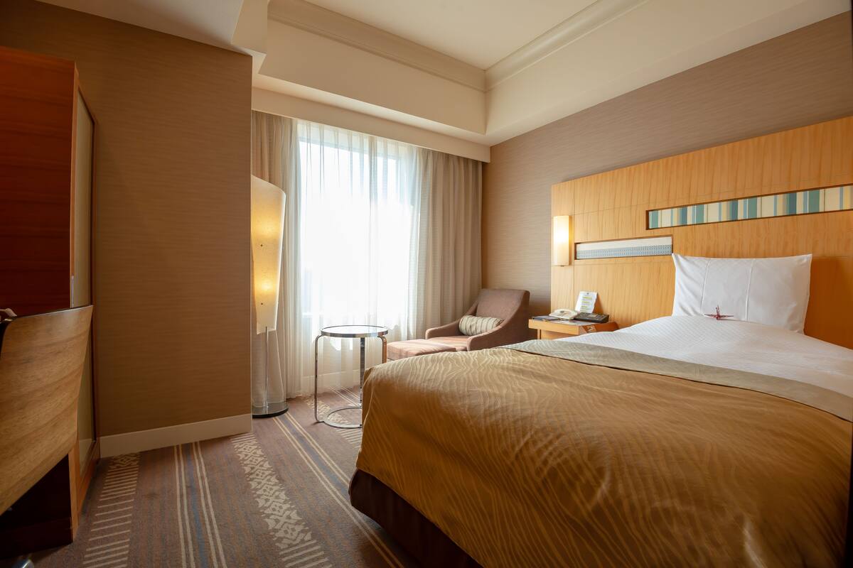 Une chambre d’hôtel avec un grand lit, une chaise, un bureau avec un téléphone et une fenêtre avec des rideaux.