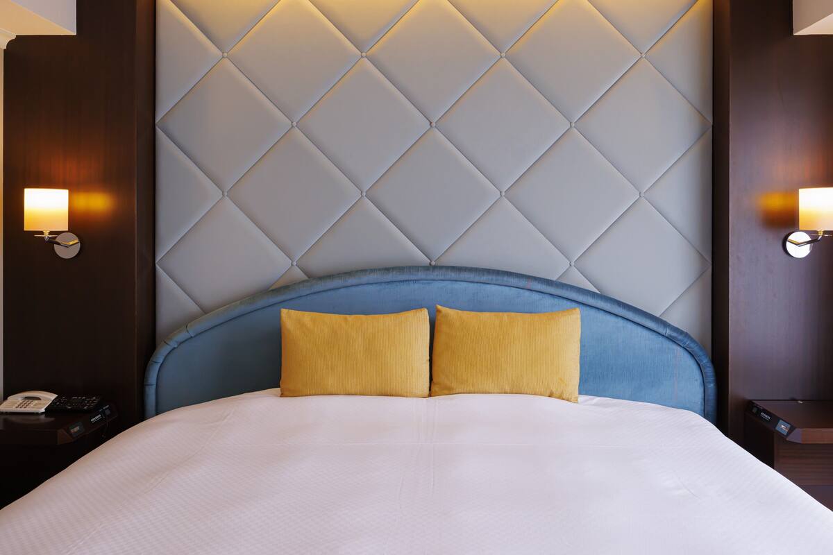 Une chambre d’hôtel avec un grand lit, une tête de lit ornée d’un motif en losanges, deux lampes fixées au mur et une table de chevet en bois avec un téléphone.