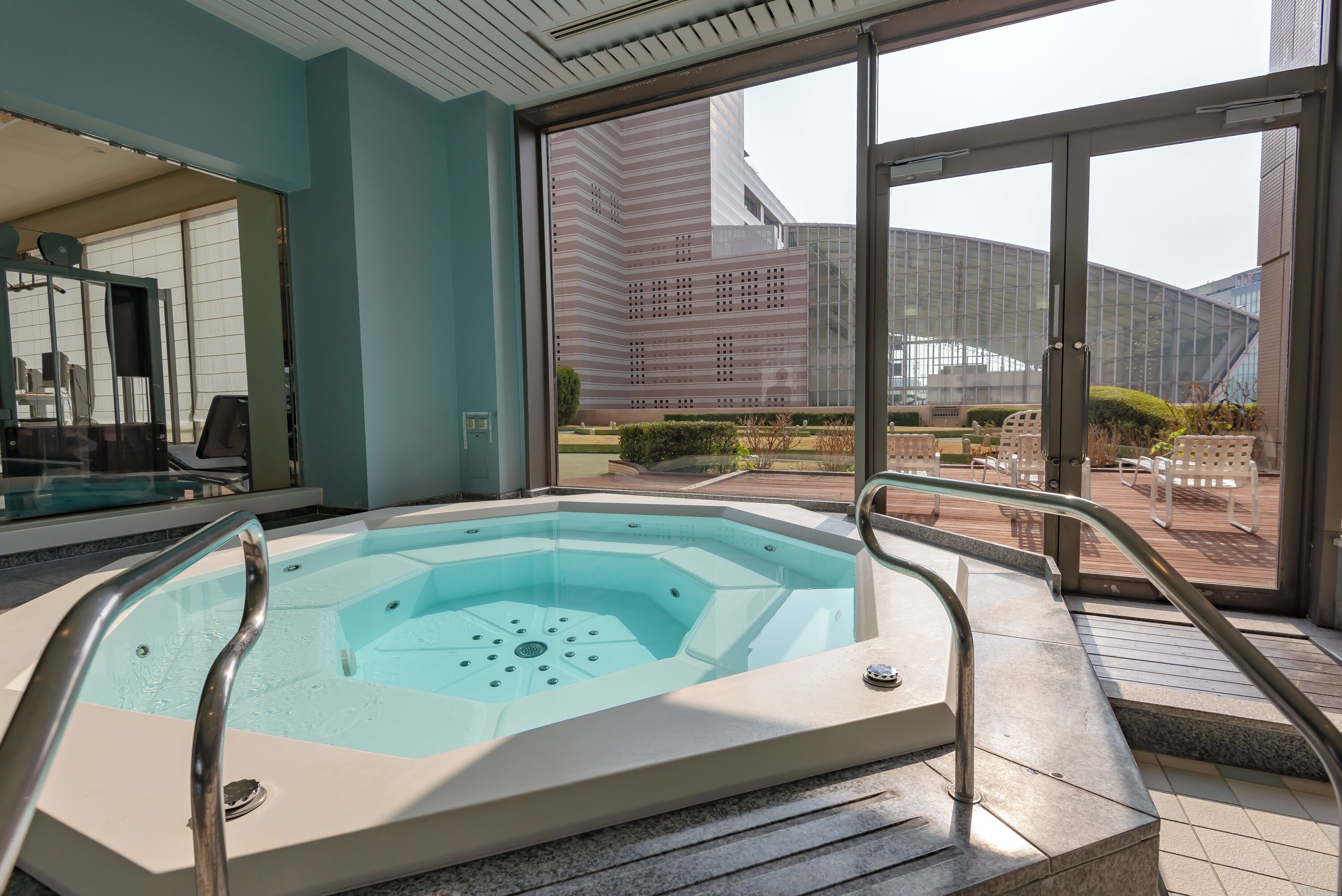 A hot tub with metal railings, a view of a modern building, and a patio area with outdoor furniture.
